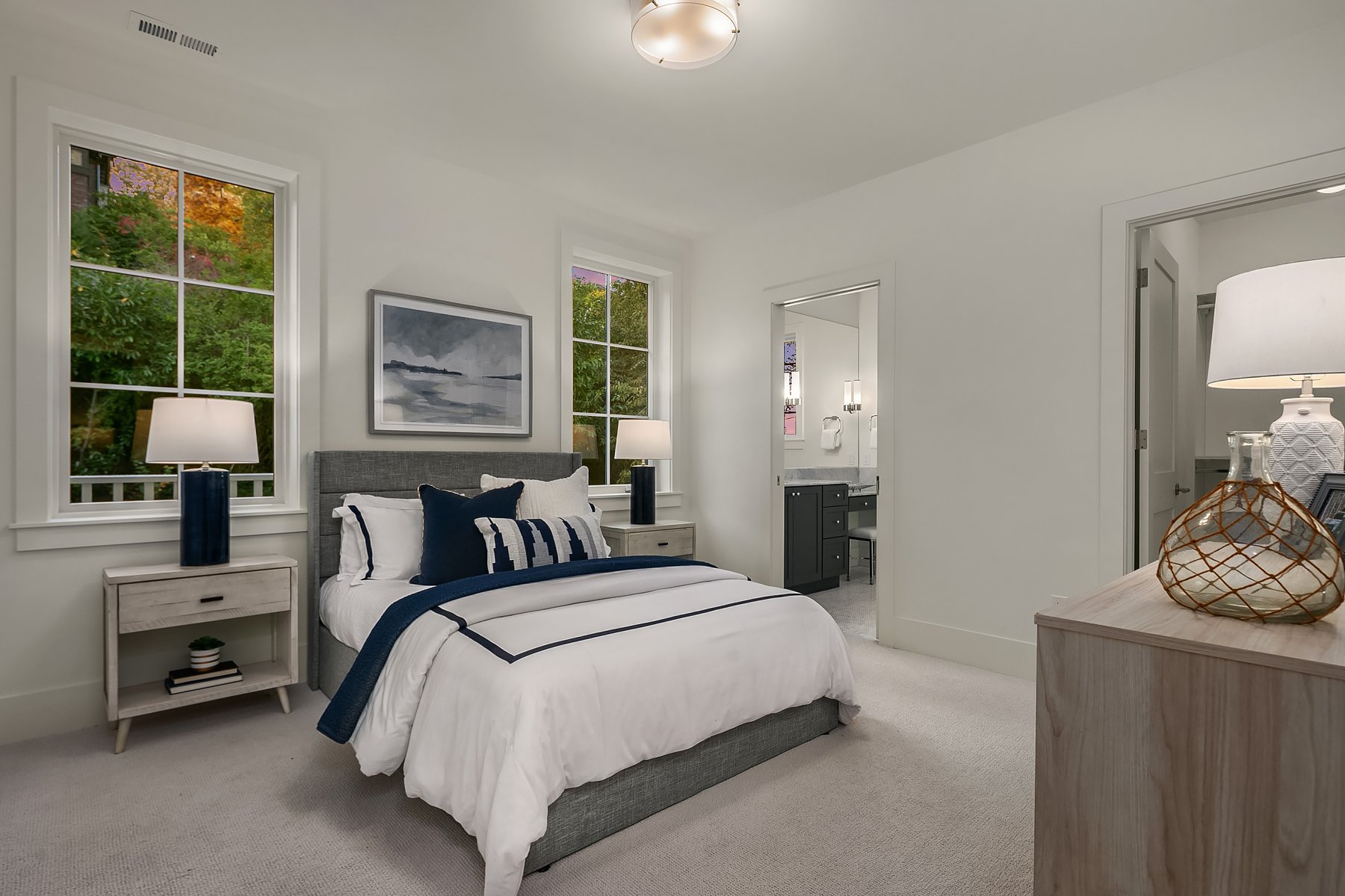 A bedroom with a gray upholstered bed, white and navy bedding, and two nightstands with lamps. There are two windows showing trees outside, with a painting above the bed. A dresser with a lamp and decorative items is visible on the right side.