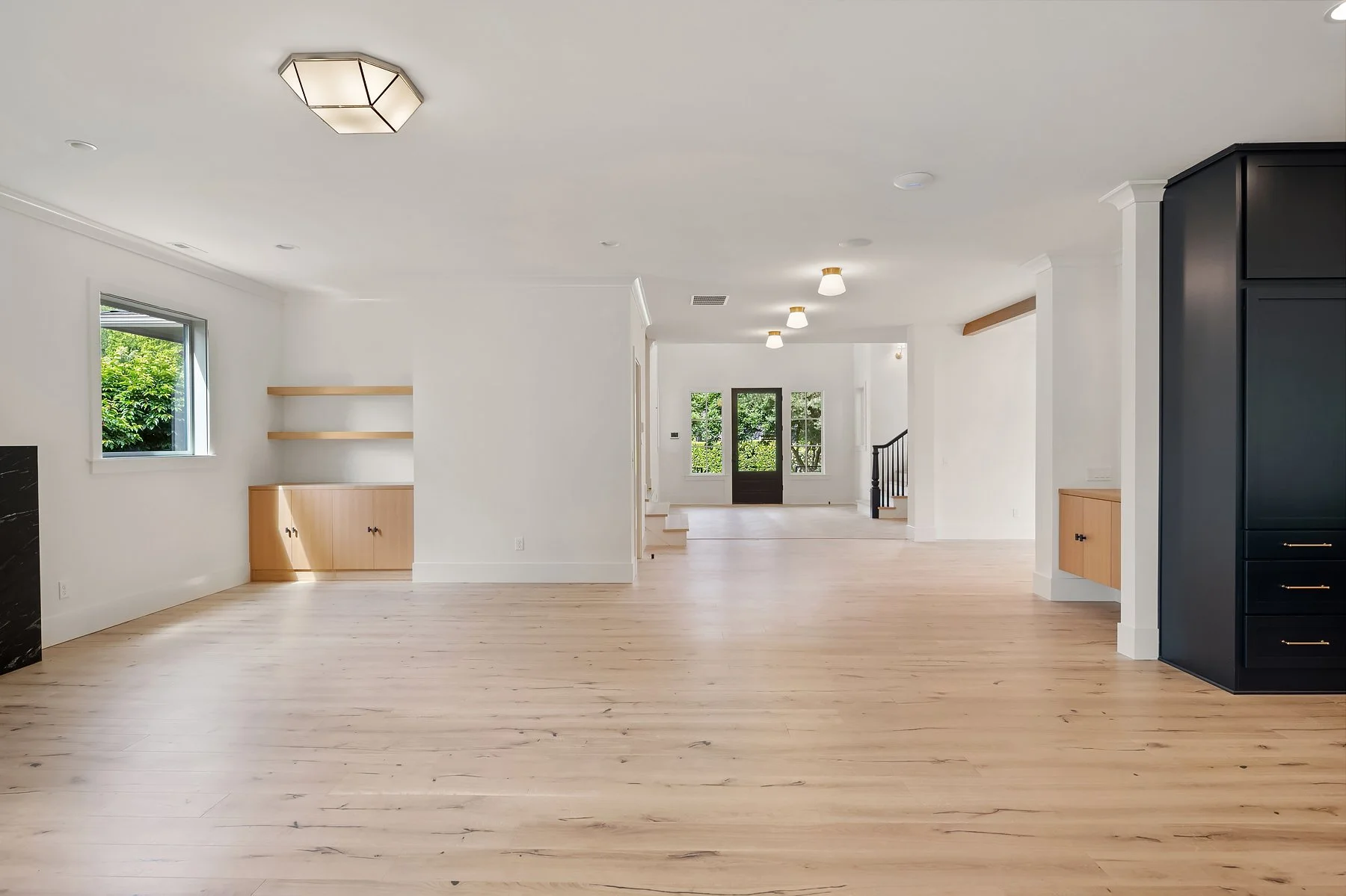 Empty living room with light wooden floors, white walls, small window showing greenery outside, built-in wooden shelves, and black cabinetry. Front door and staircase visible in the background.