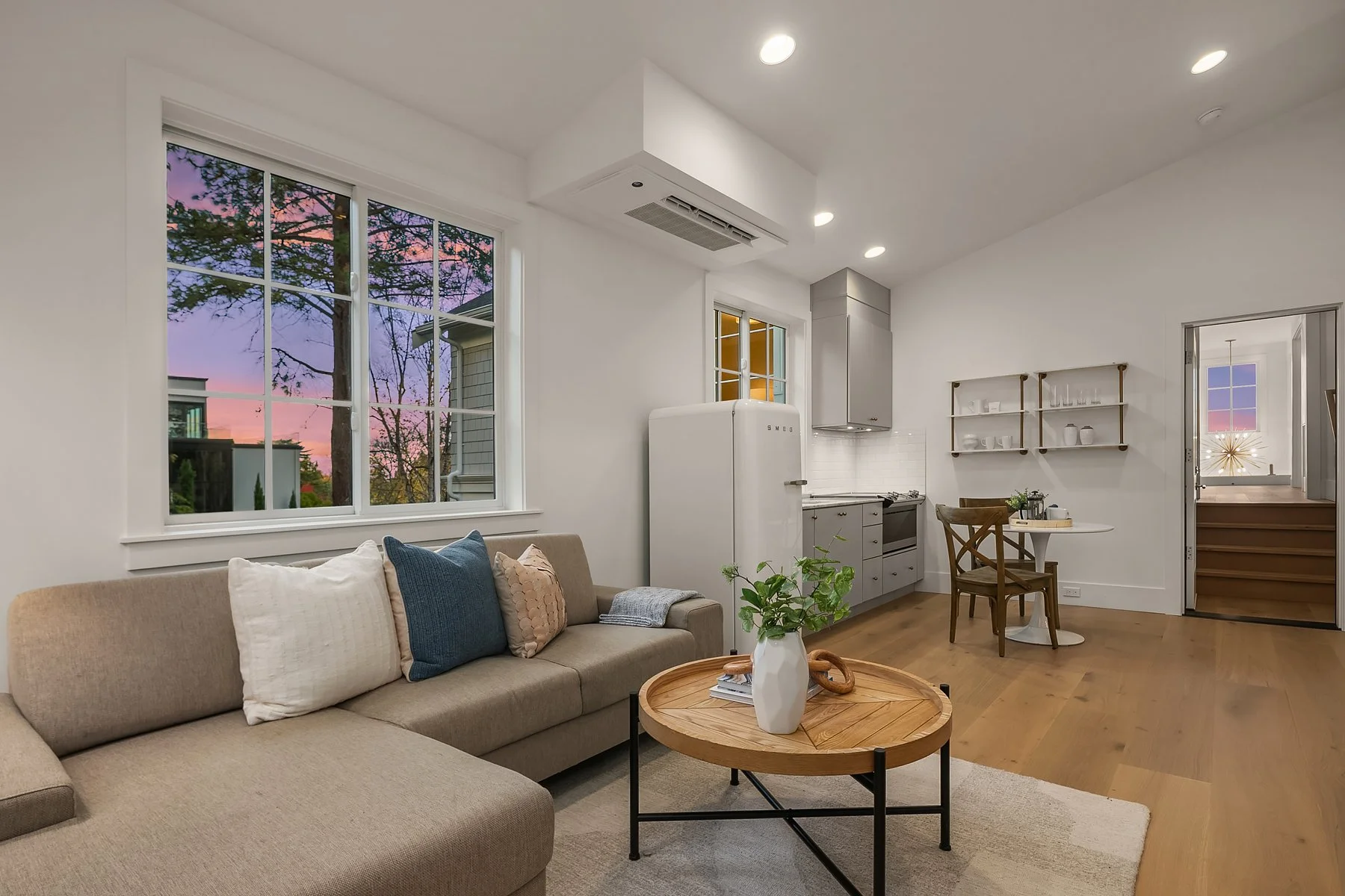 Cozy living room with beige sectional sofa, colorful pillows, round wooden coffee table with a plant and magazines, large window showing sunset sky and trees, open kitchen with gray cabinets, vintage refrigerator, small dining table with chair, and a