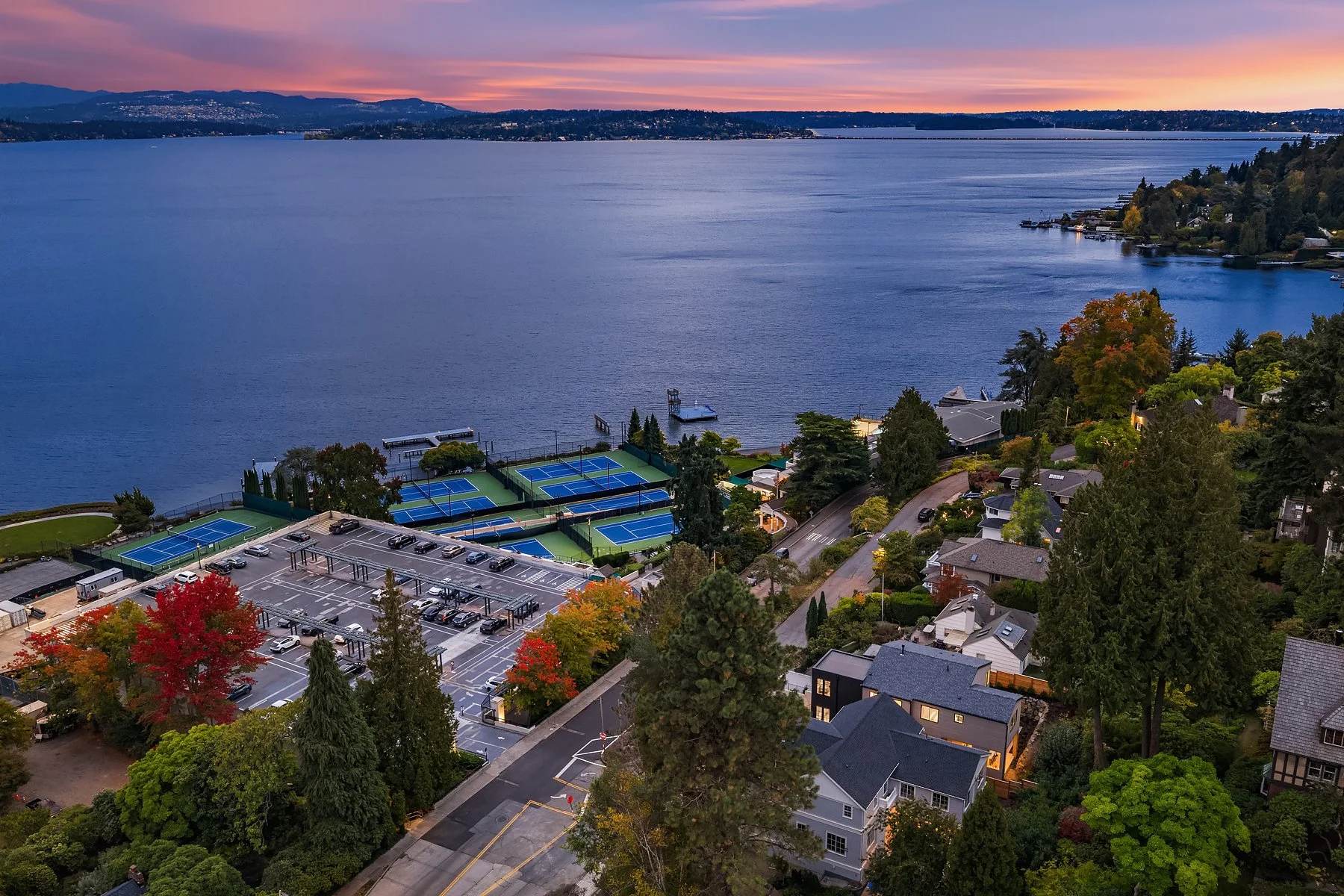 Aerial view of a residential neighborhood with a lake and tennis courts, showing autumn trees with red, orange, and green leaves at sunset.
