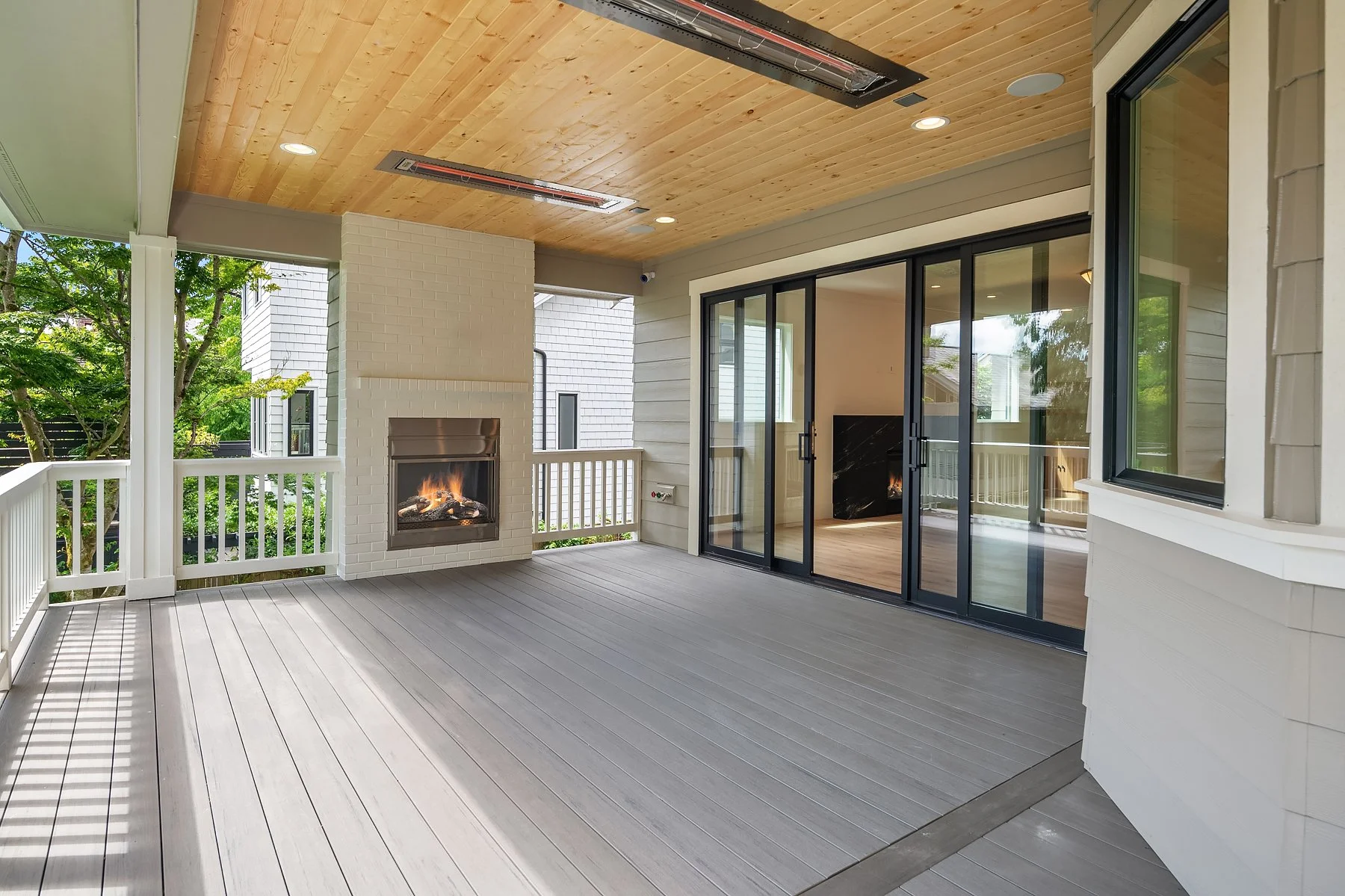 Covered outdoor patio with fireplace, sliding glass doors, and wood ceiling, overlooking green trees outside.