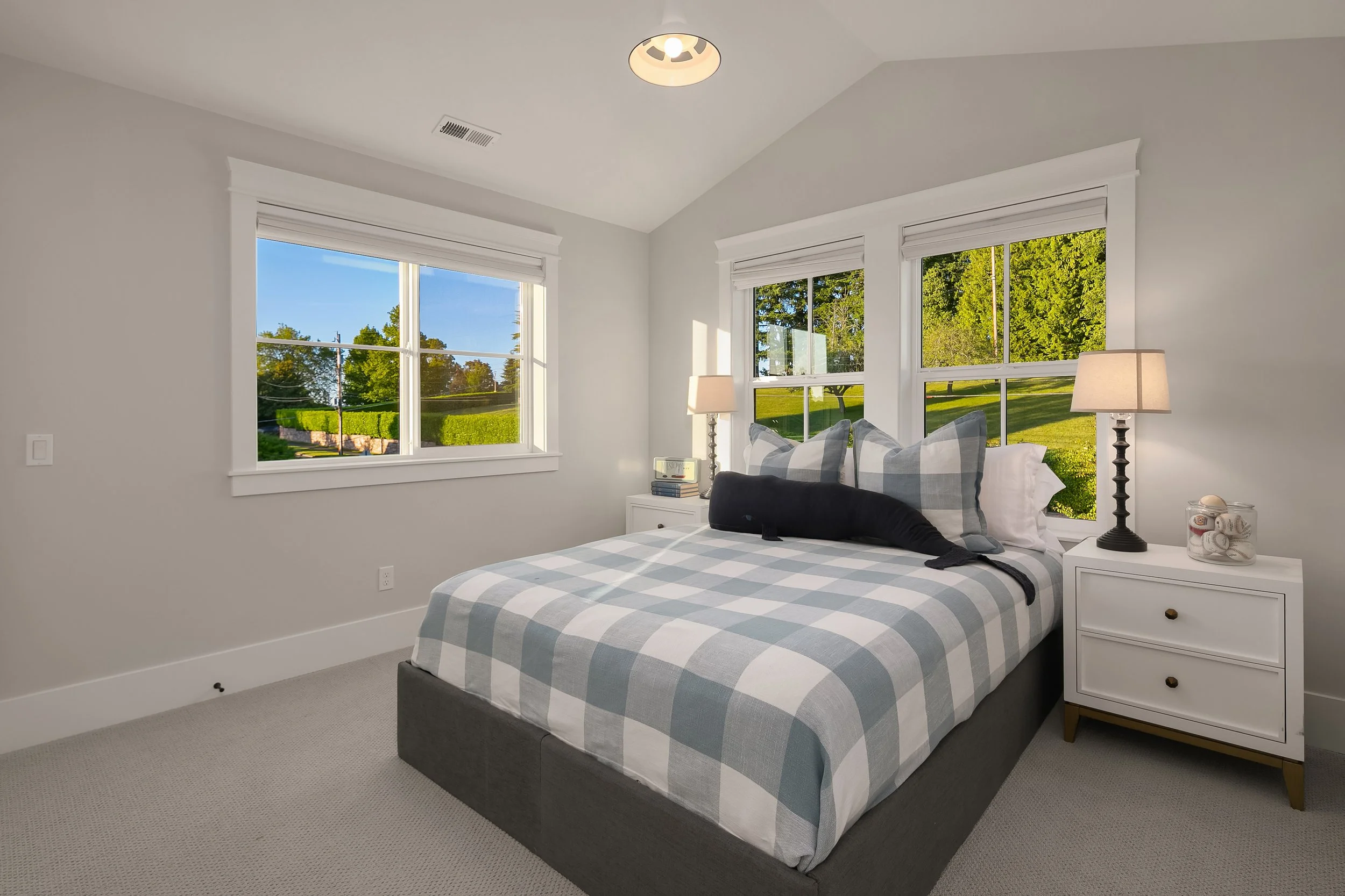 Bright bedroom with two large windows showing green trees and grass outside. The bed has plaid bedding in shades of gray and white, with multiple pillows. There are two white nightstands with lamps, one on each side of the bed. One nightstand has a j