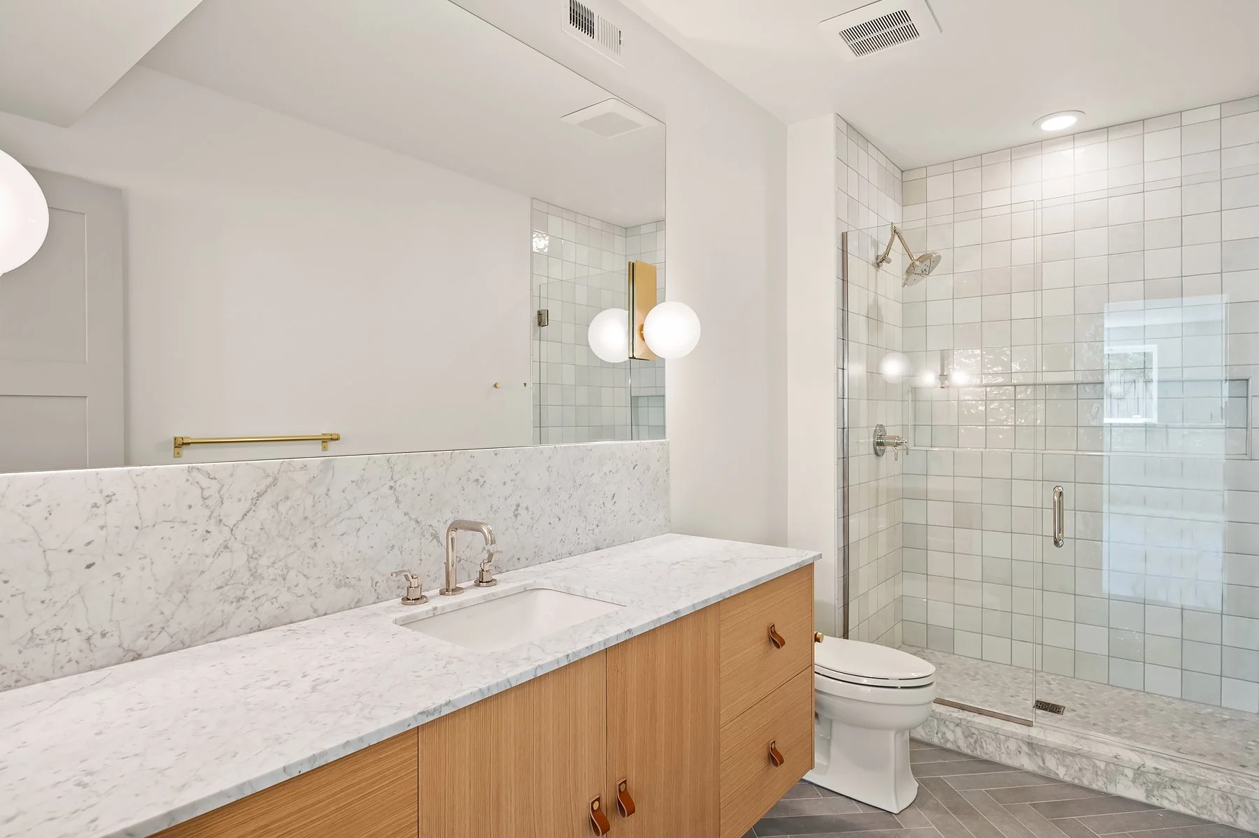 Modern bathroom with a wooden vanity with a marble countertop, a white sink, a mirror, wall-mounted light fixtures, a toilet, and a glass-enclosed shower with white tile walls and a marble base.