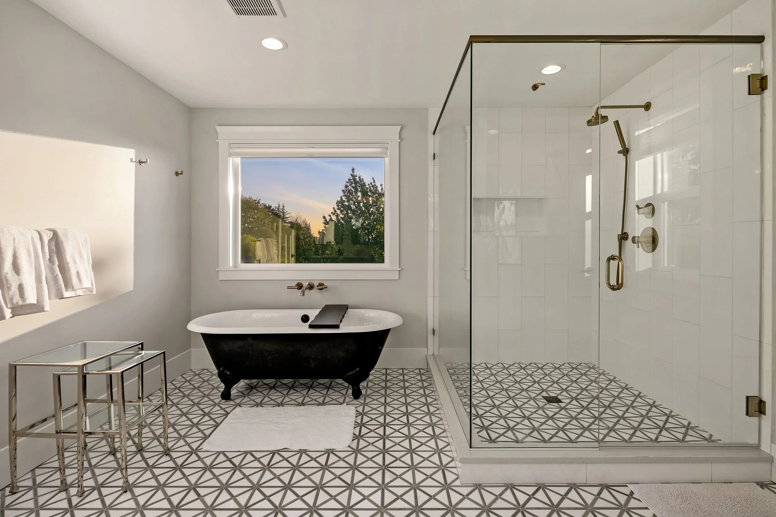 Modern bathroom with black clawfoot tub, glass-enclosed shower, white tiled walls, patterned floor tiles, window showing greenery outside, and minimalist decor.