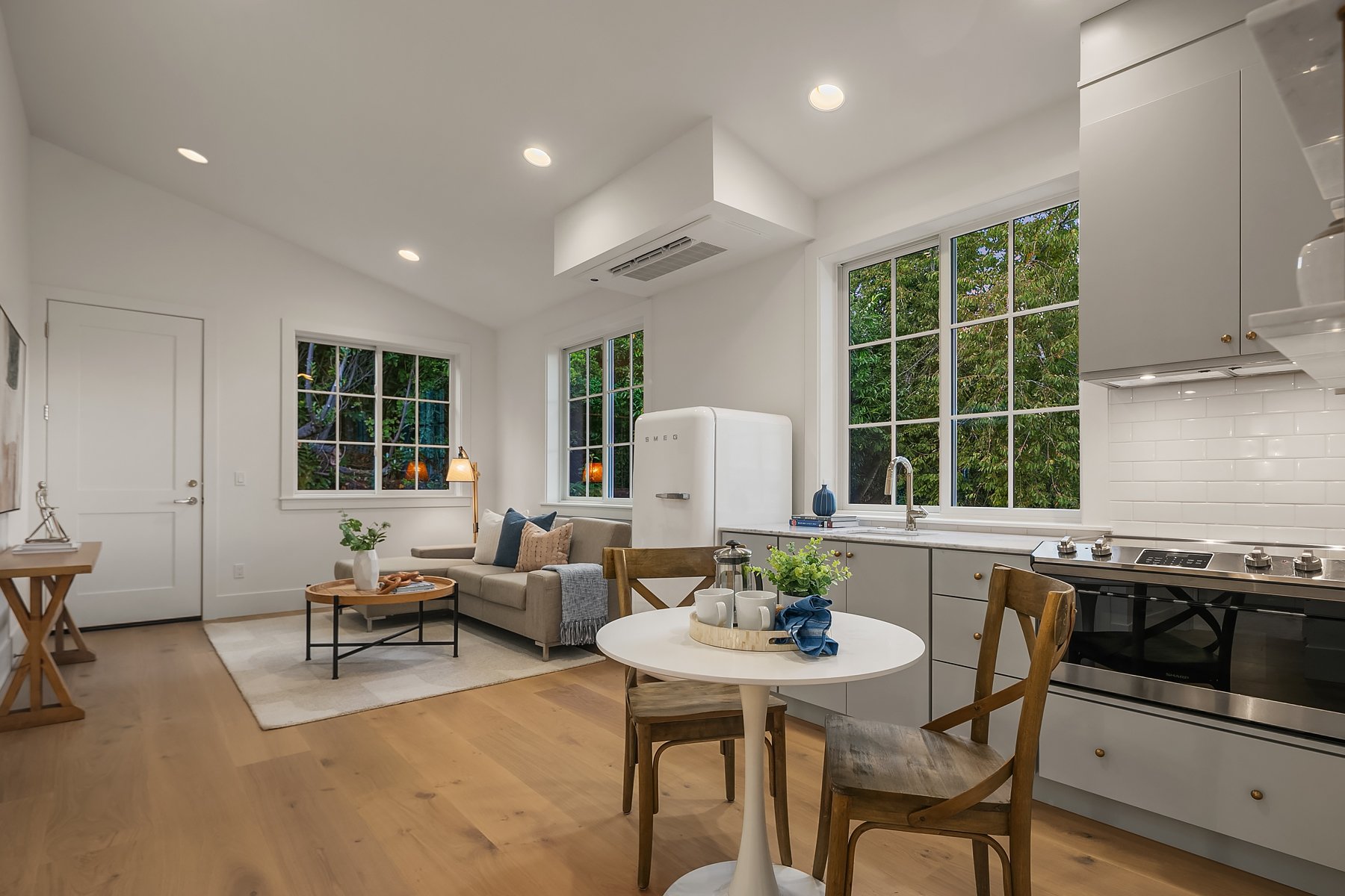 Bright open-concept kitchen and living room with white walls, hardwood floors, large windows, gray cabinets, and minimalist decor.