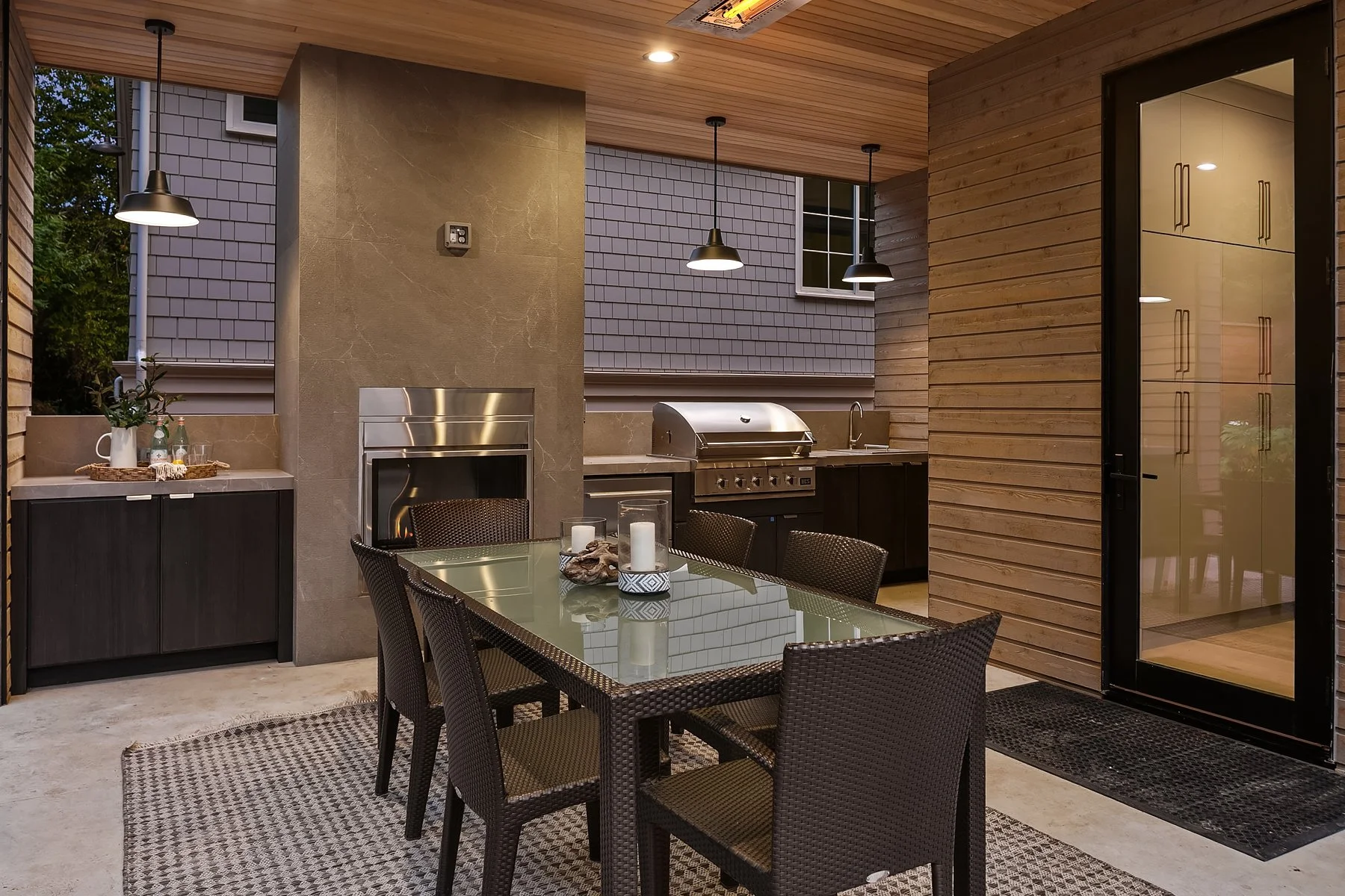 Outdoor patio with a dining table and chairs, grill, fireplace, and kitchen area, enclosed by wood and glass walls, with ceiling lights