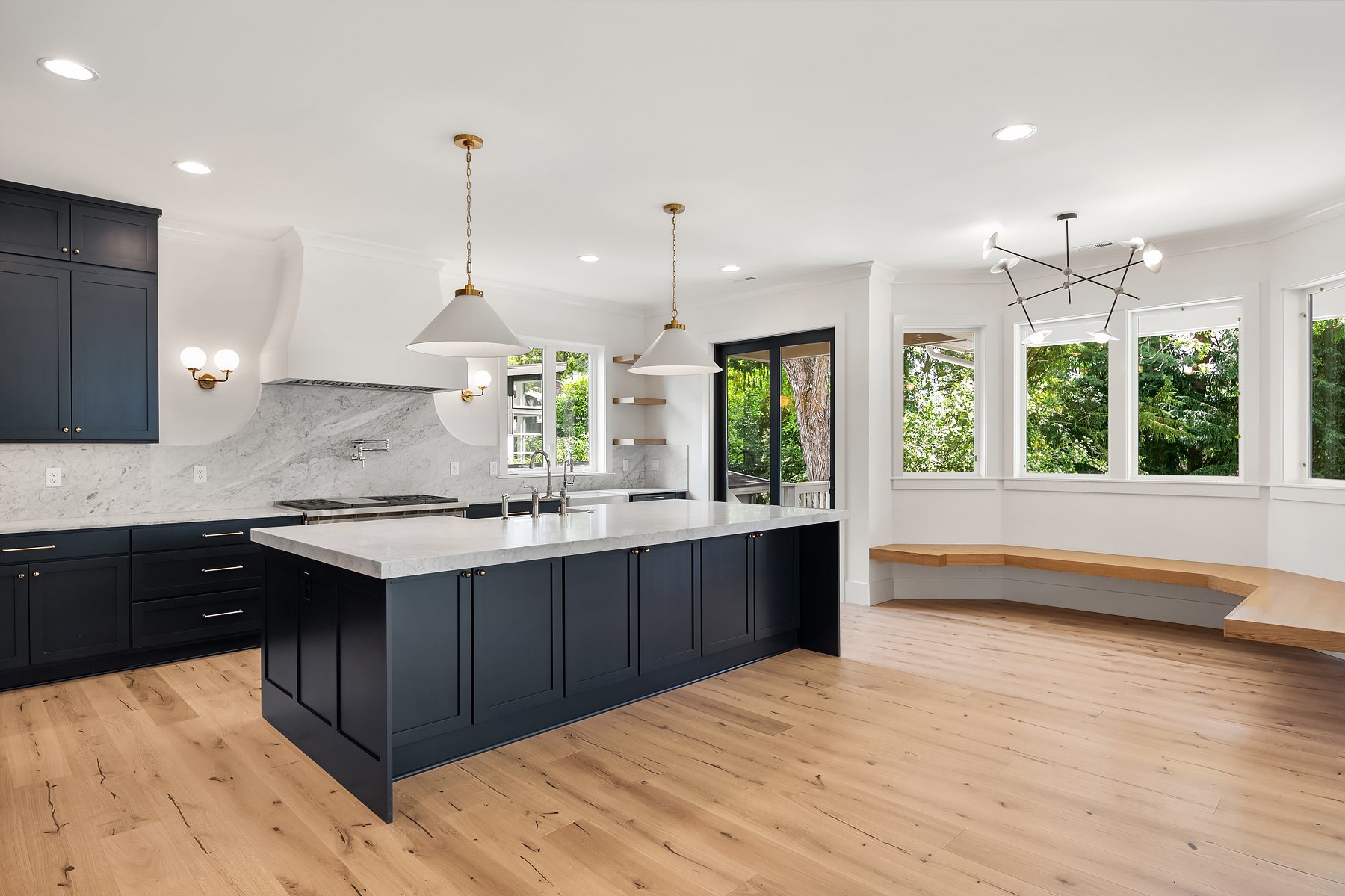 Modern kitchen with navy blue cabinets, white marble countertops, a large island, light wood flooring, and large windows with views of greenery.