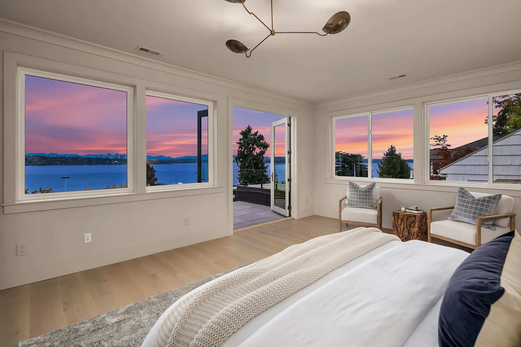 Bedroom with large windows showing a colorful sunset over a lake, with a door leading to a balcony, and a bed with white bedding and navy blue pillows, along with two armchairs and a wooden side table.