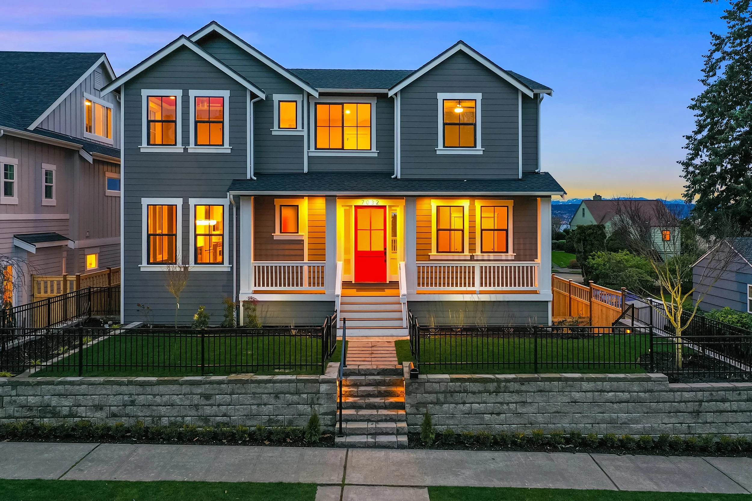 A two-story house with gray siding, white trim, and a red front door, illuminated from within, located in a suburban neighborhood during twilight.
