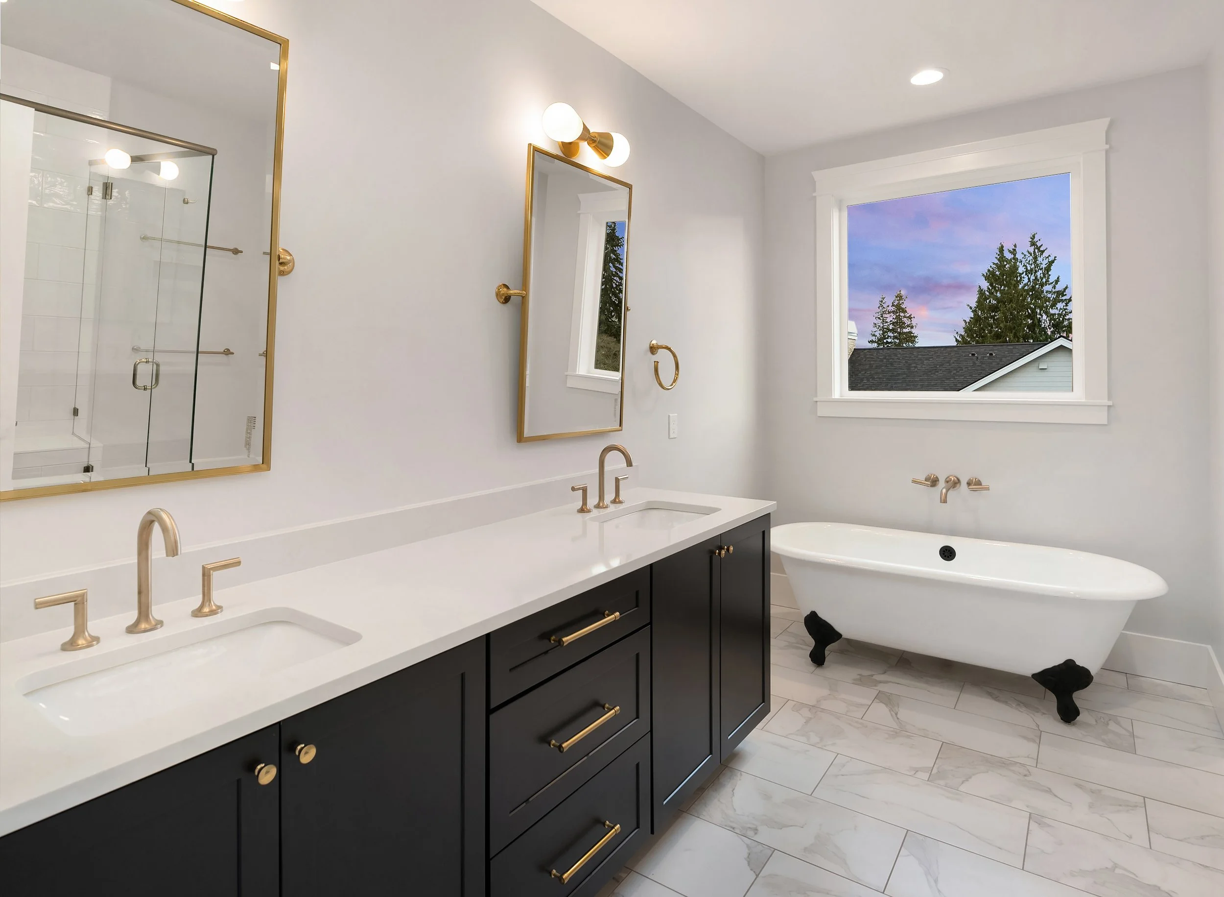 Modern bathroom with double vanity, gold fixtures, and a freestanding clawfoot bathtub near a window showing an evening sky.