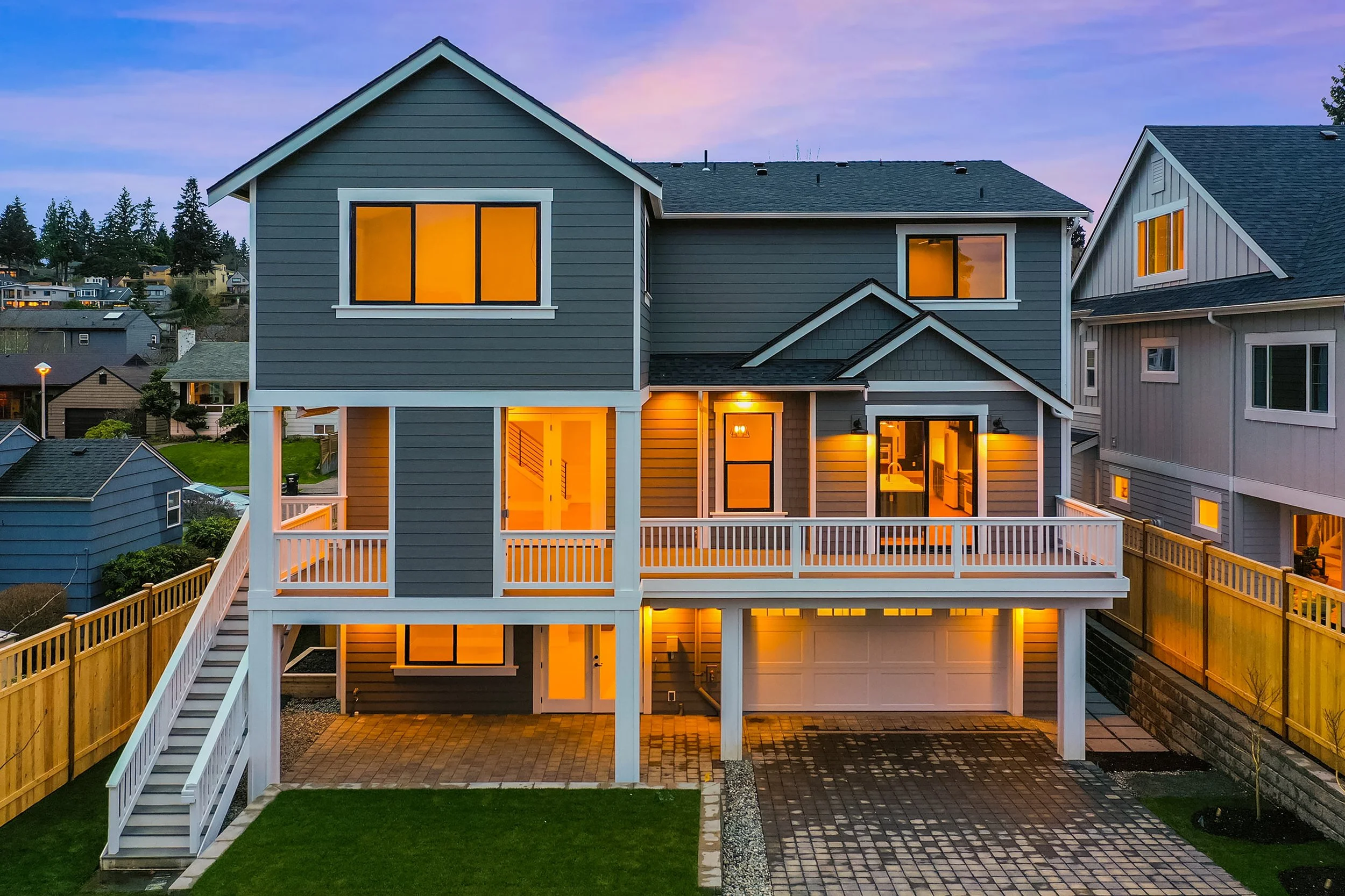 A modern, multi-story house with grey siding and white trim, illuminated from inside, on a slightly elevated lot with a paved driveway and a wooden staircase leading to a front porch, surrounded by a wooden fence.
