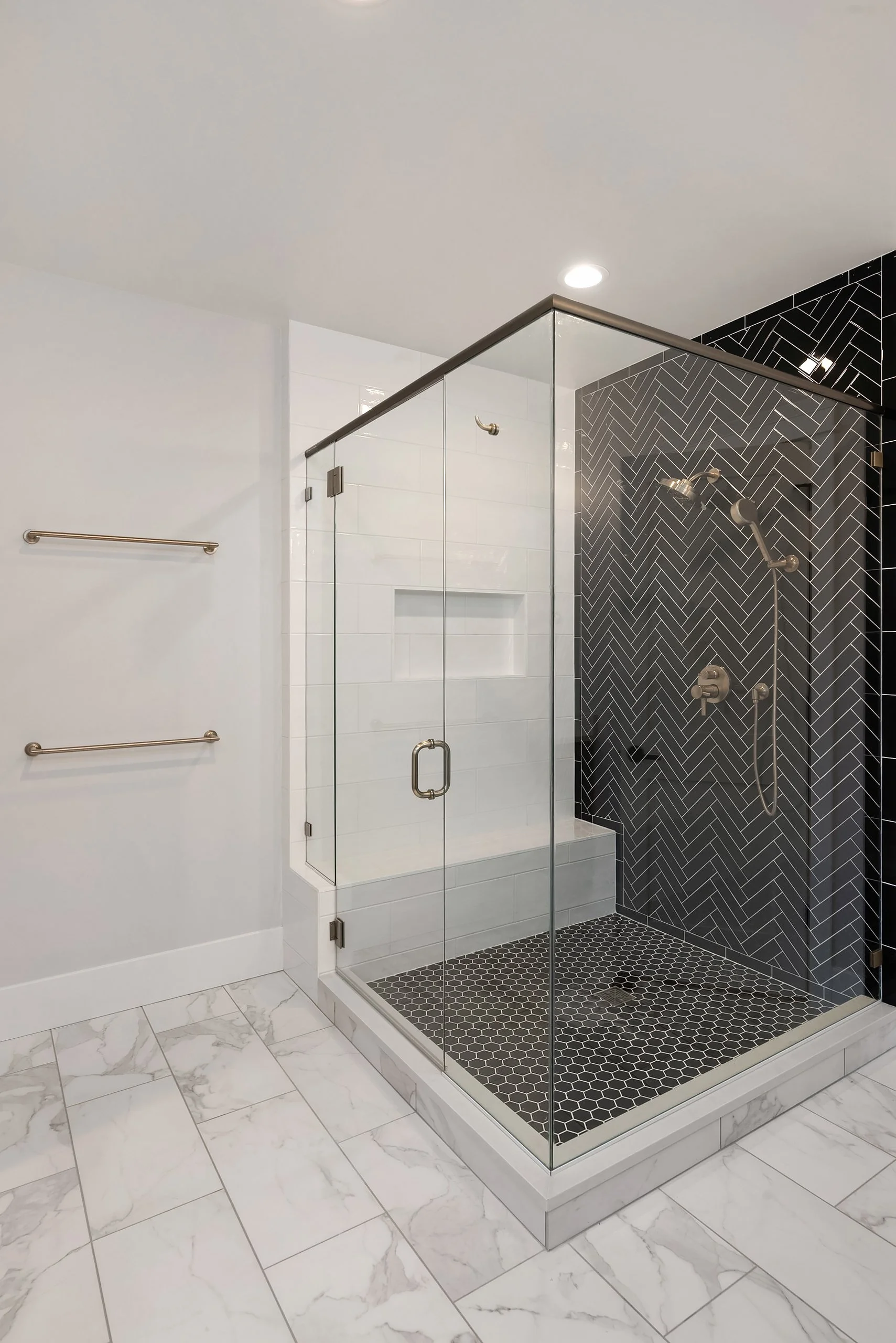 Modern corner shower with clear glass doors, black and white herringbone and hex tile accents, and white marble flooring in a bathroom.