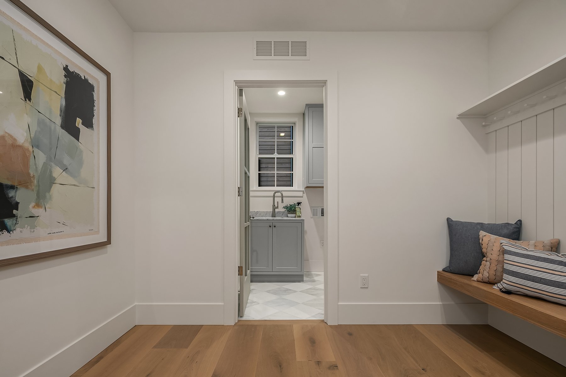 Interior view of a room with a doorway leading to a small kitchenette with a window, gray cabinets, and a porcelain sink. The room has white walls, hardwood flooring, a wooden bench with pillows, and a large abstract painting on the left wall.