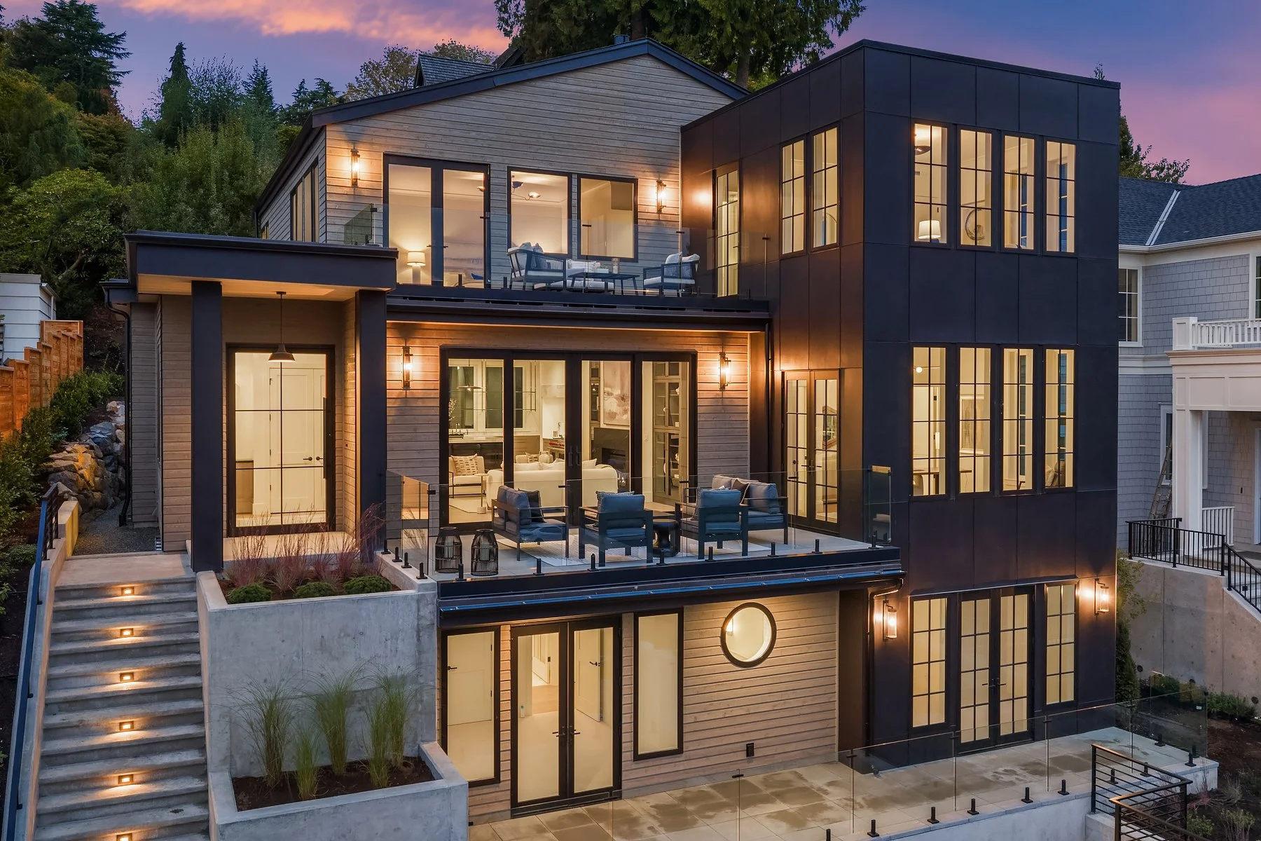 Modern multi-story house with large glass windows and outdoor seating areas, situated on a hillside with landscaping and outdoor lighting at dusk.