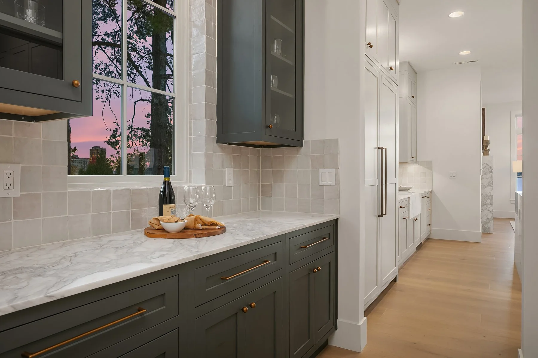A modern kitchen with gray and white cabinets, marble countertops, and a window showing a sunset sky with trees.