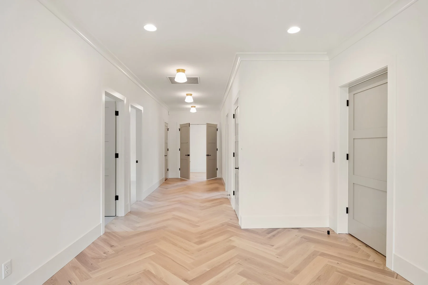 Empty hallway with white walls, light wood chevron flooring, ceiling lights, and multiple closed doors.