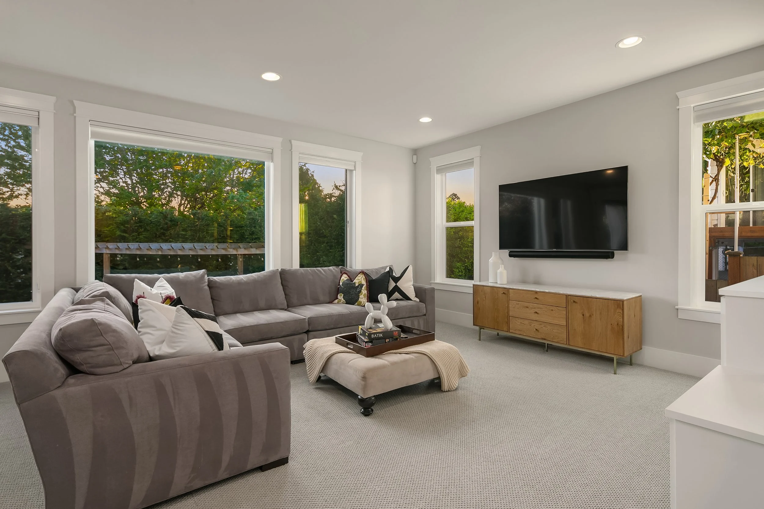 Living room with gray sectional sofa, large windows showing green trees outside, modern wooden TV console, mounted flat-screen TV, decorative items on the console, cream-colored carpet, and white walls with crown molding.