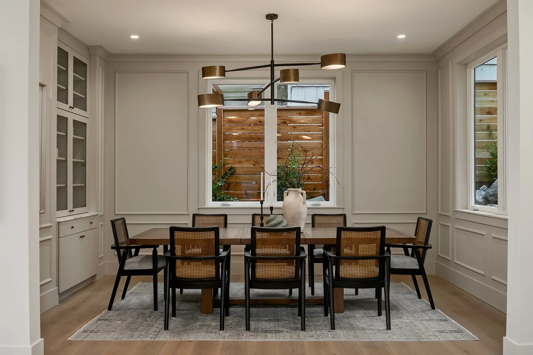 Modern dining room with a wooden table, nine chairs, a large vase with branches and greenery, wood-paneled fence visible outside through the window, white paneled walls, built-in cabinets, and a contemporary chandelier.