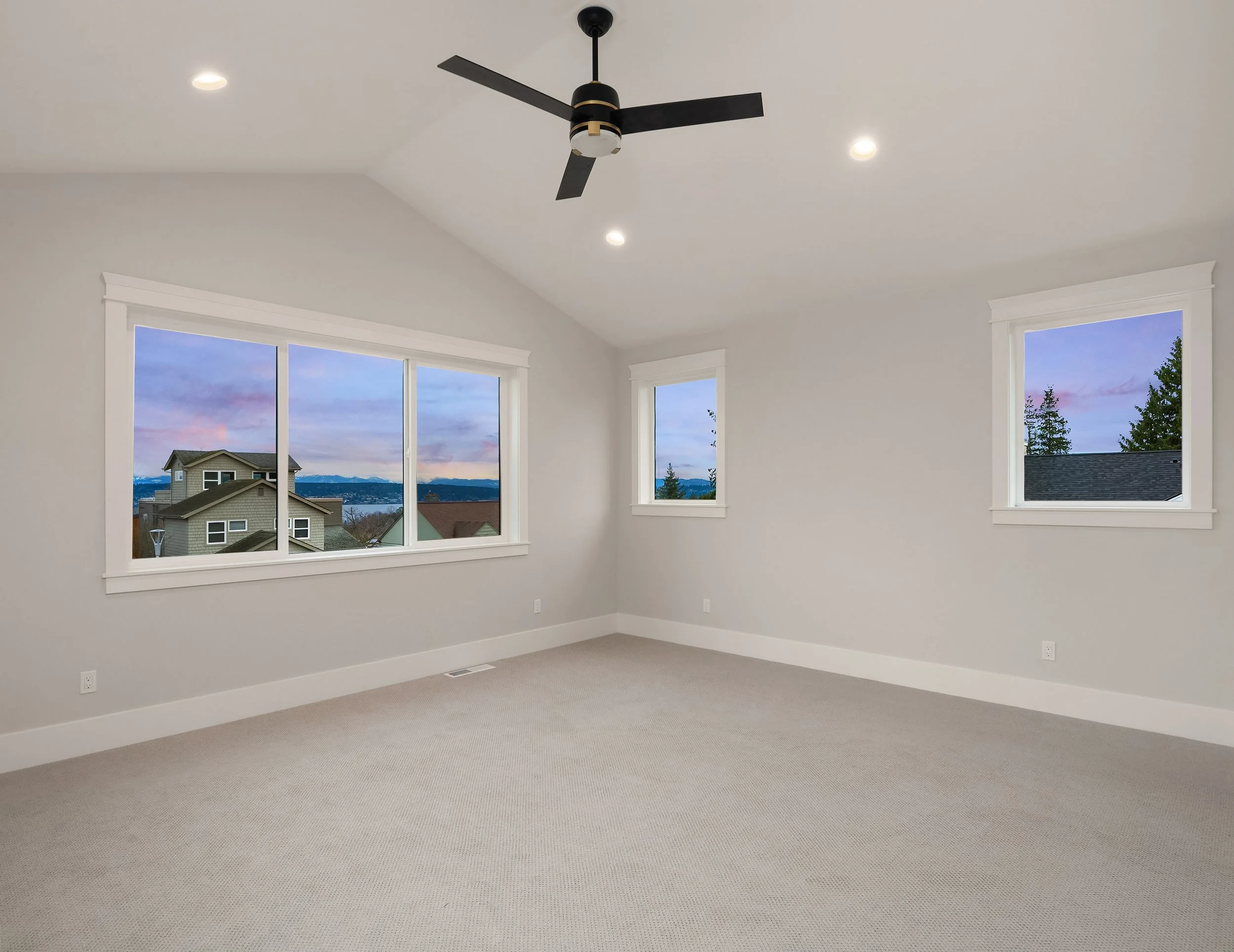 Empty room with three large windows showing a neighborhood and mountains, ceiling fan, recessed lighting, light-colored walls, carpeted floor.