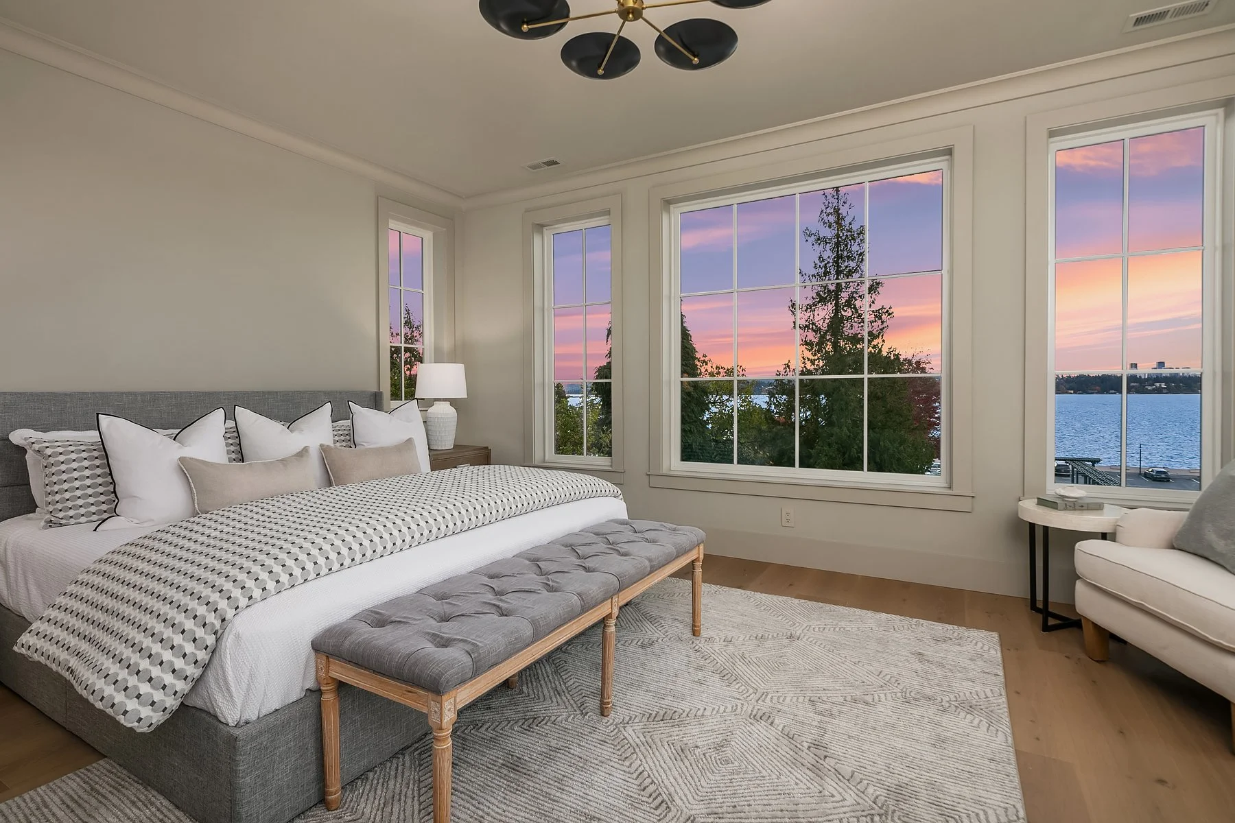 Bedroom with large windows showing a sunset over a body of water, with a bed, pillows, a bench, and a side table with a lamp.