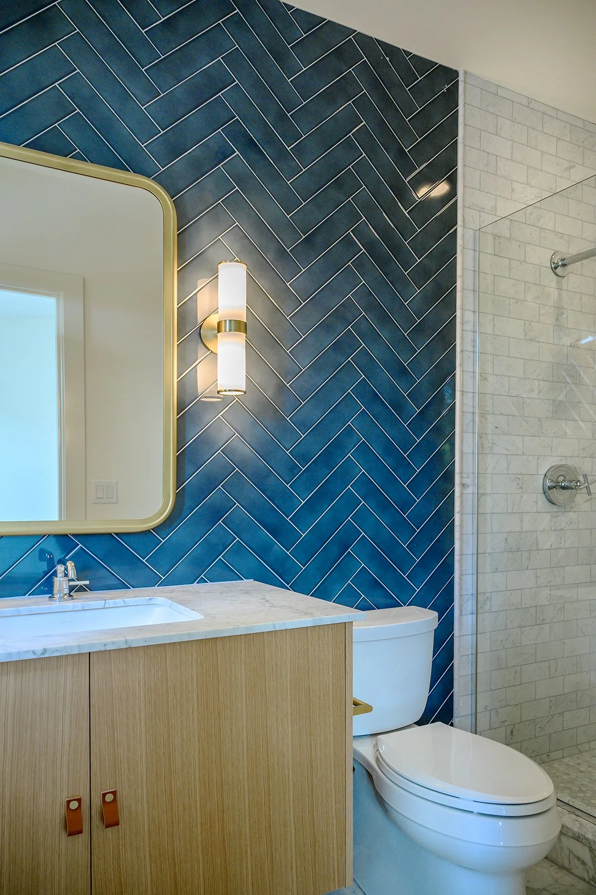 Modern bathroom with blue herringbone tiled wall, white toilet, wooden vanity with marble countertop, and shower with white tiles.