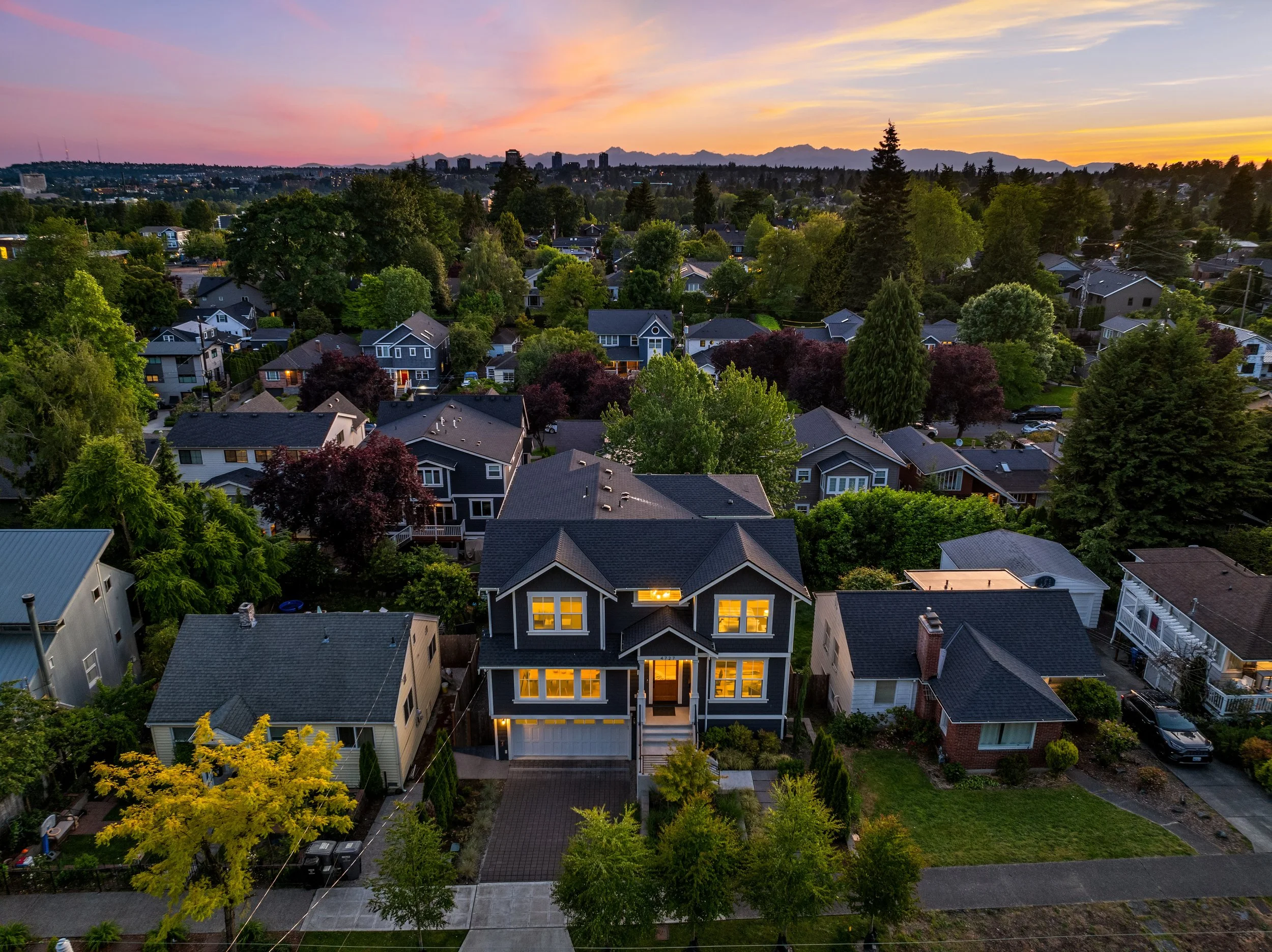 Aerial view of a suburban neighborhood at sunset showing a large, illuminated house with a dark roof and multiple lit windows, surrounded by green trees and other houses.