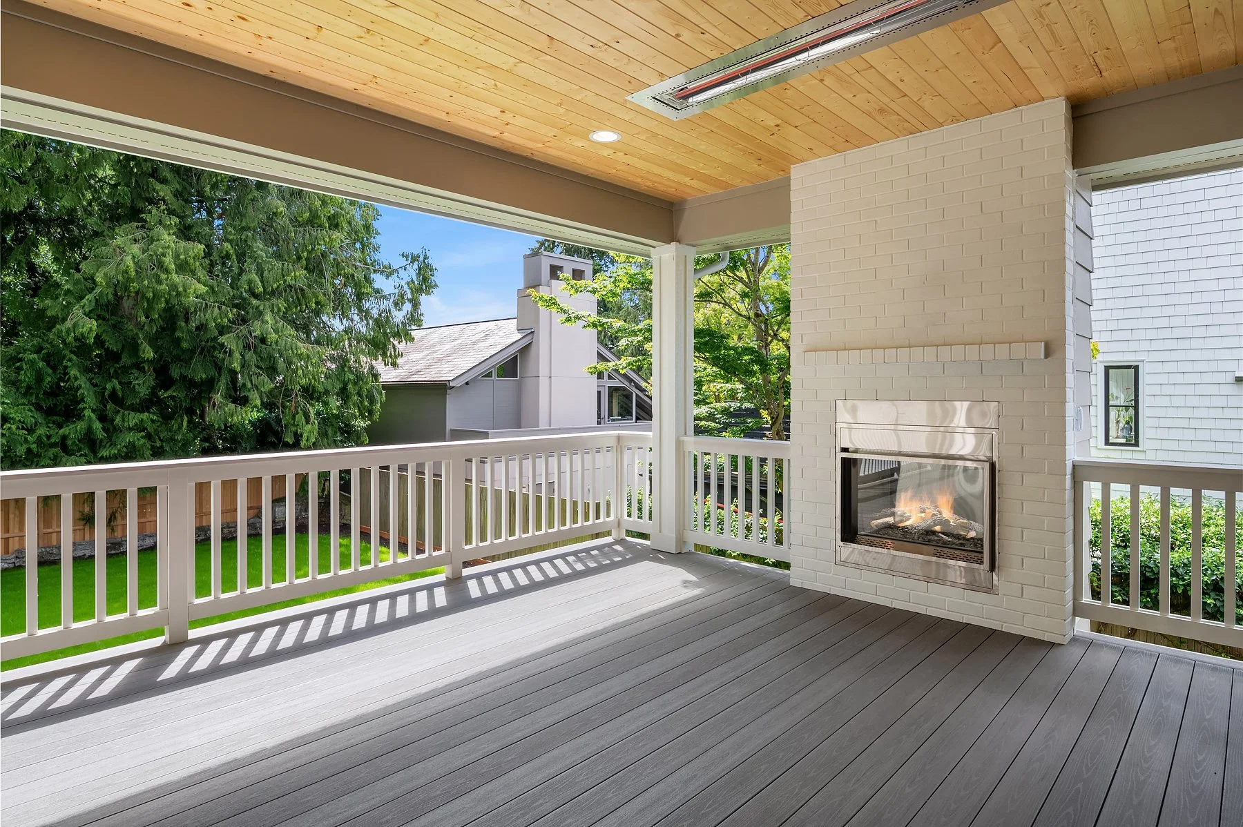 Covered outdoor porch with a built-in fireplace, white railing, gray floorboards, wood-paneled ceiling, and a view of neighboring house and trees.