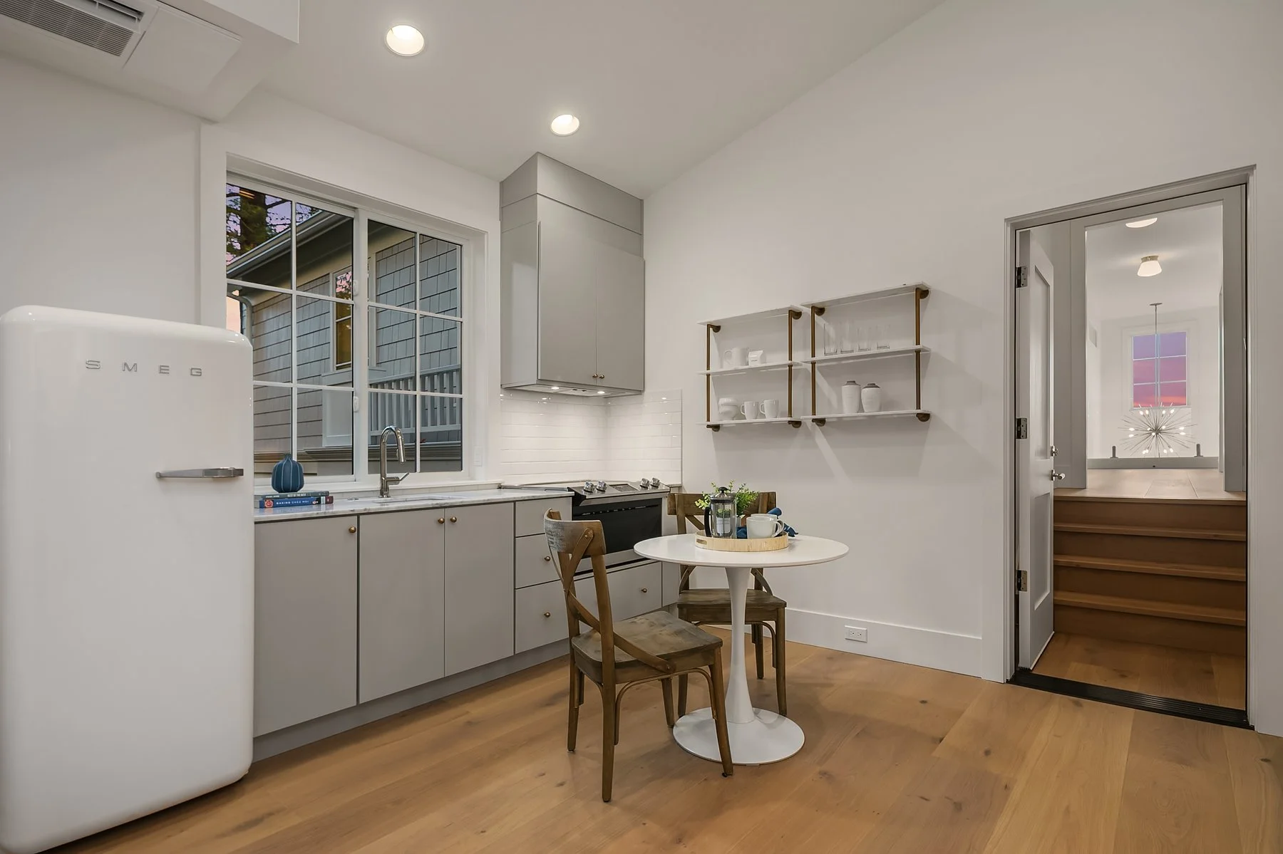 A modern kitchen with light gray cabinets, a white backsplash, and a window above the sink. There is a white Smeg refrigerator on the left, a small round white dining table with two wooden chairs in the center, and open white shelves on the wall. The