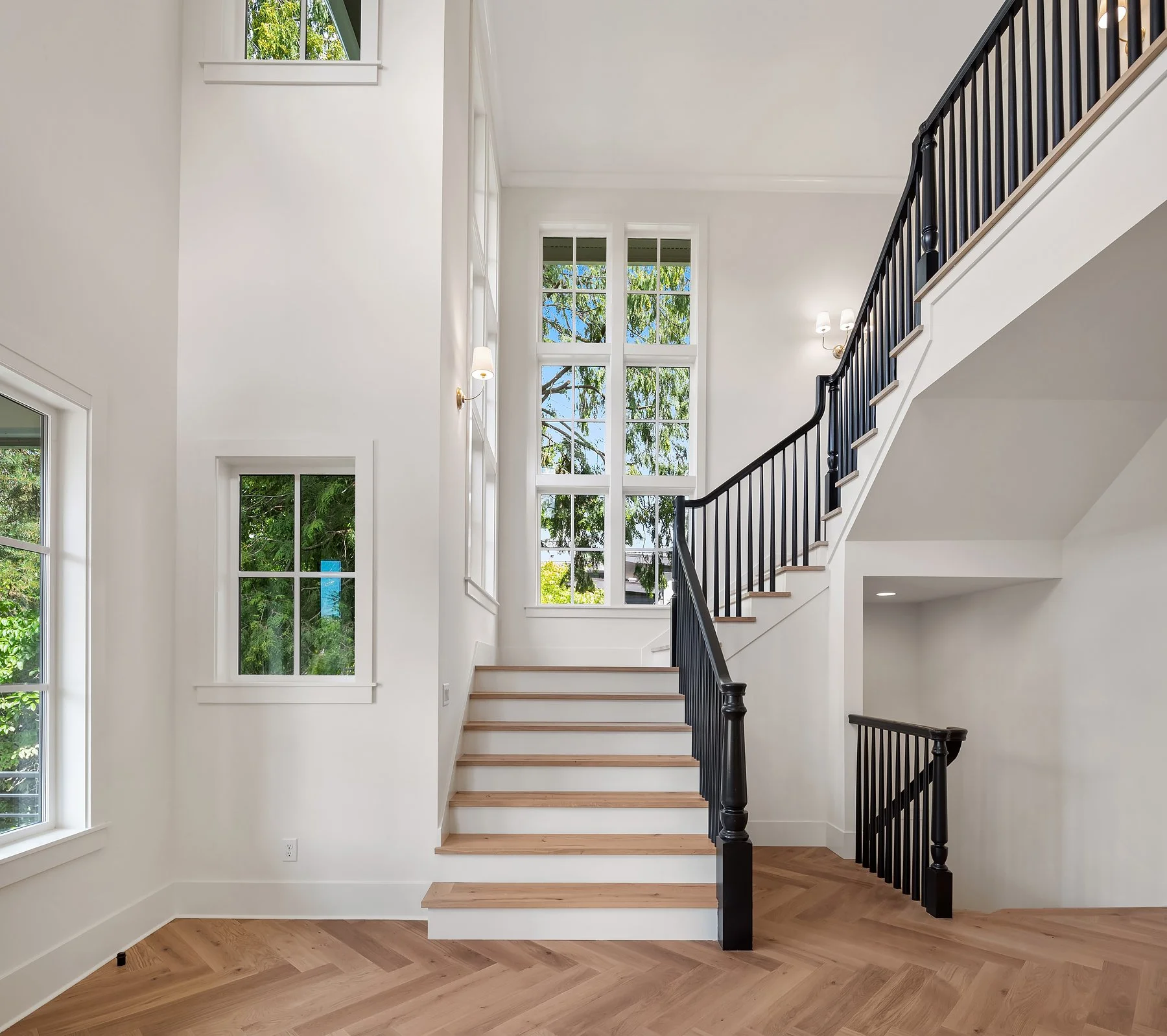 Interior view of a modern home with white walls, hardwood floors, large multi-pane windows, and a staircase with black railing.