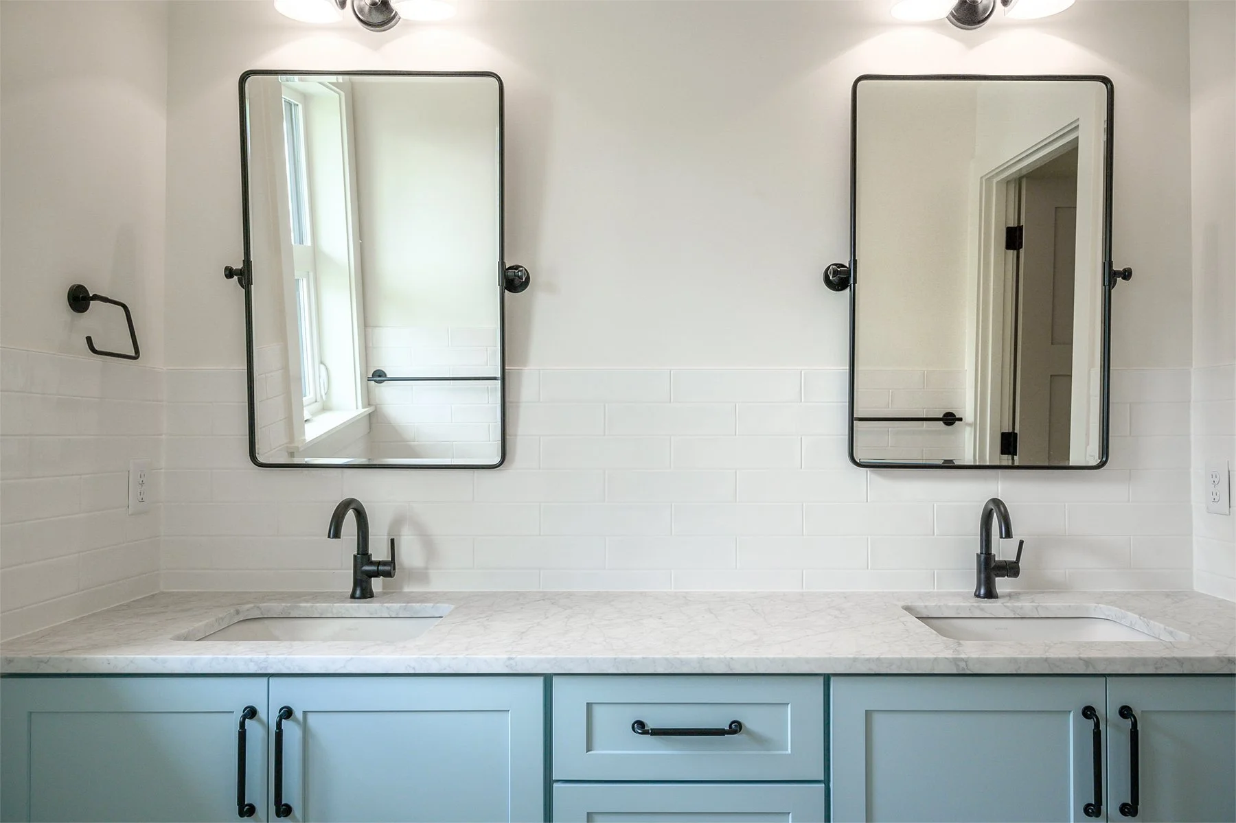 Double vanity bathroom with two rectangular mirrors, black faucets, light blue cabinets, white marble countertop, white subway tile backsplash, and wall-mounted light fixtures.