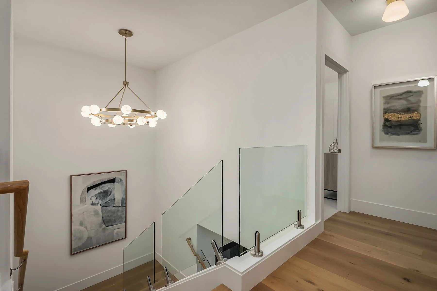 Modern staircase landing with wooden flooring, abstract artwork on white walls, glass balustrades, and a contemporary chandelier.