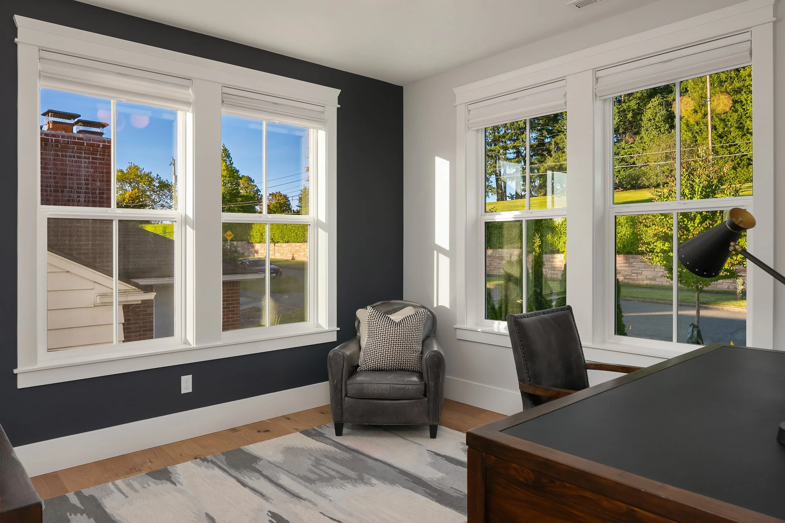 A corner of a modern living room with three large windows showing a yard with trees and a street. There is a grey armchair with a black and white cushion, a black desk lamp, and part of a wooden desk.