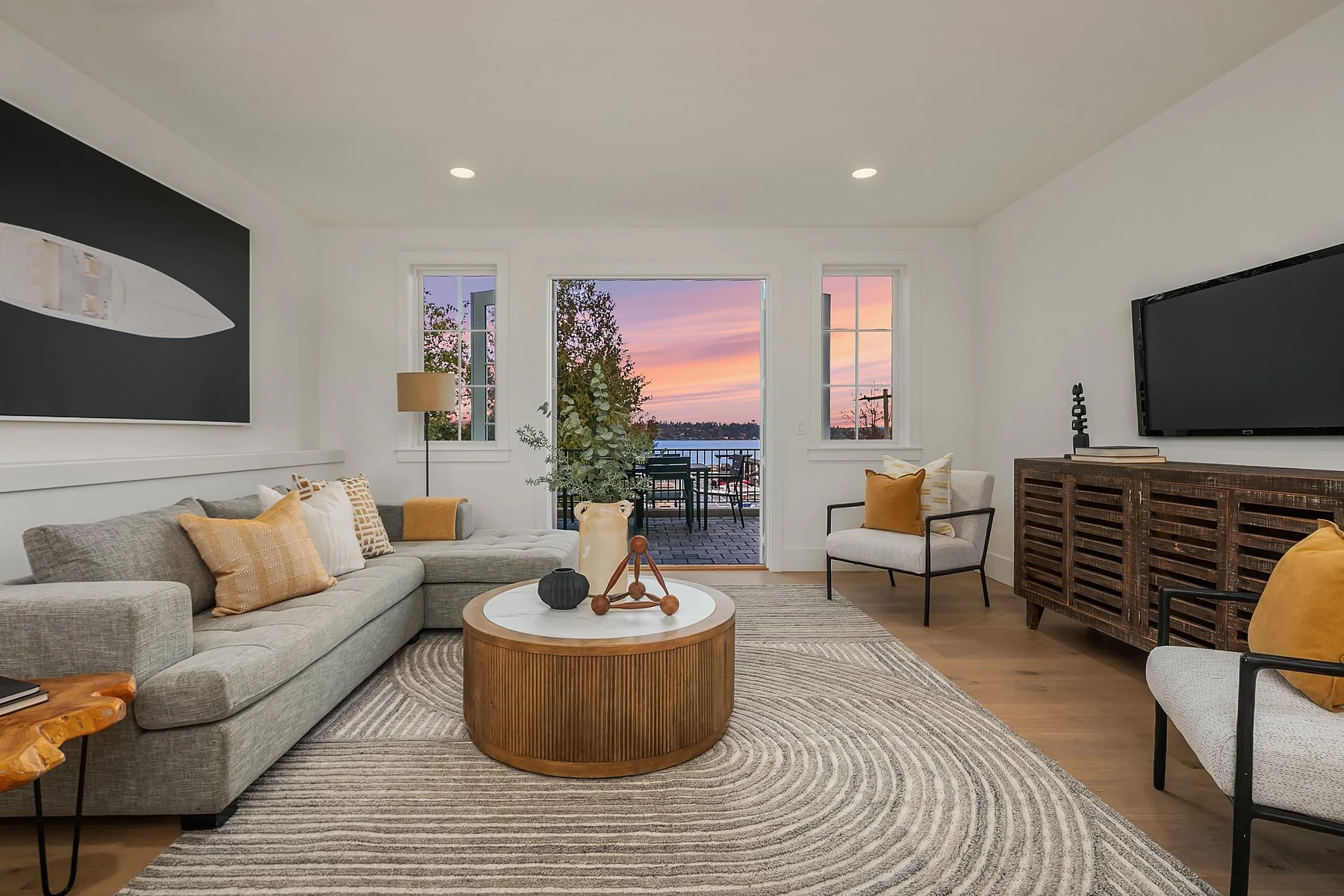 Living room with a large gray sectional sofa, a round wooden coffee table, and two white armchairs with mustard pillows. There is a flat-screen TV on a wooden media console, and a door leading to an outdoor balcony with a view of the sunset over wate