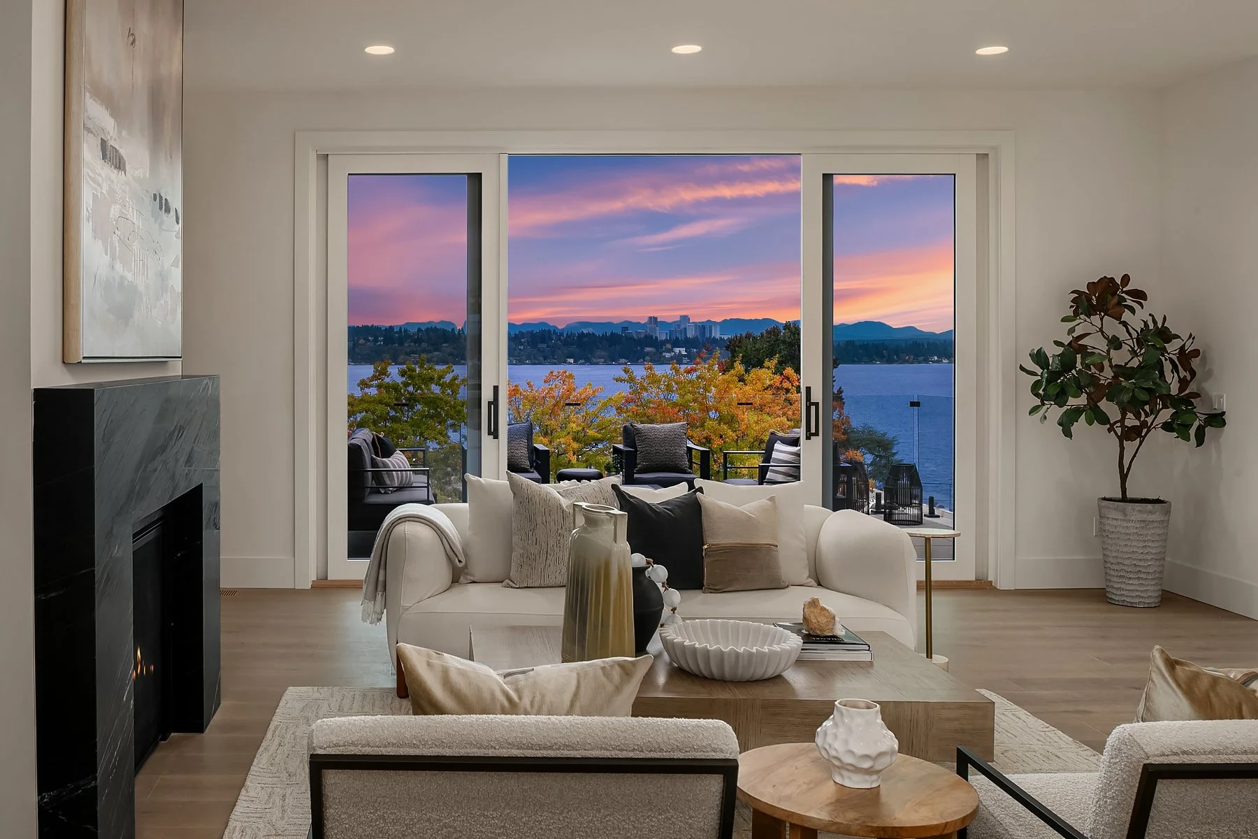 Living room with white sofa, decorative pillows, artwork on wall, wooden coffee table with vases and decorative objects, sliding glass door opening to balcony with outdoor chairs, view of lake, trees with autumn foliage, city skyline, and mountains a