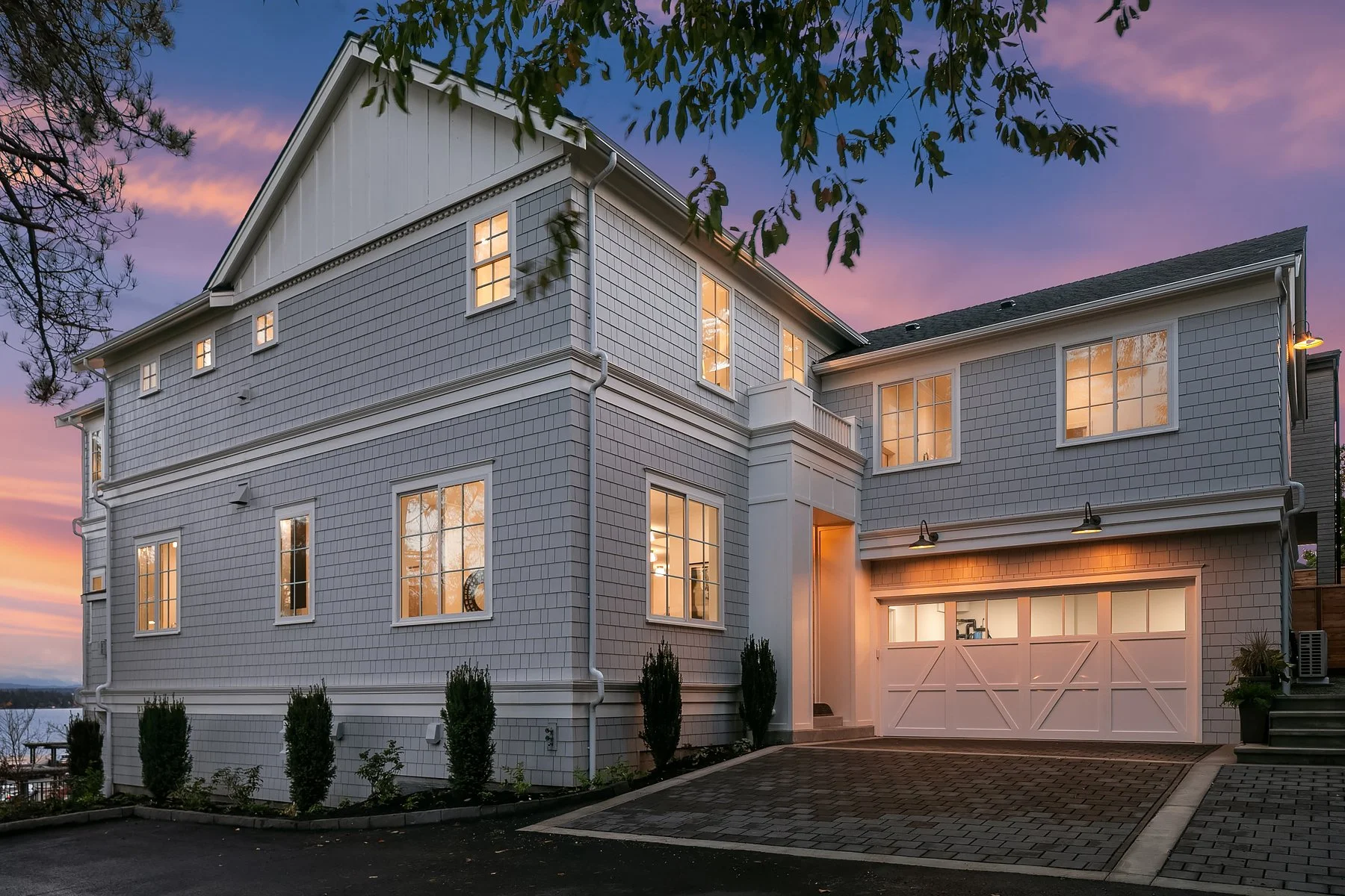 Modern multi-story house with white siding and large windows illuminated from inside, evening sky with purple and pink hues, driveway leading to garage, landscaped bushes in front.