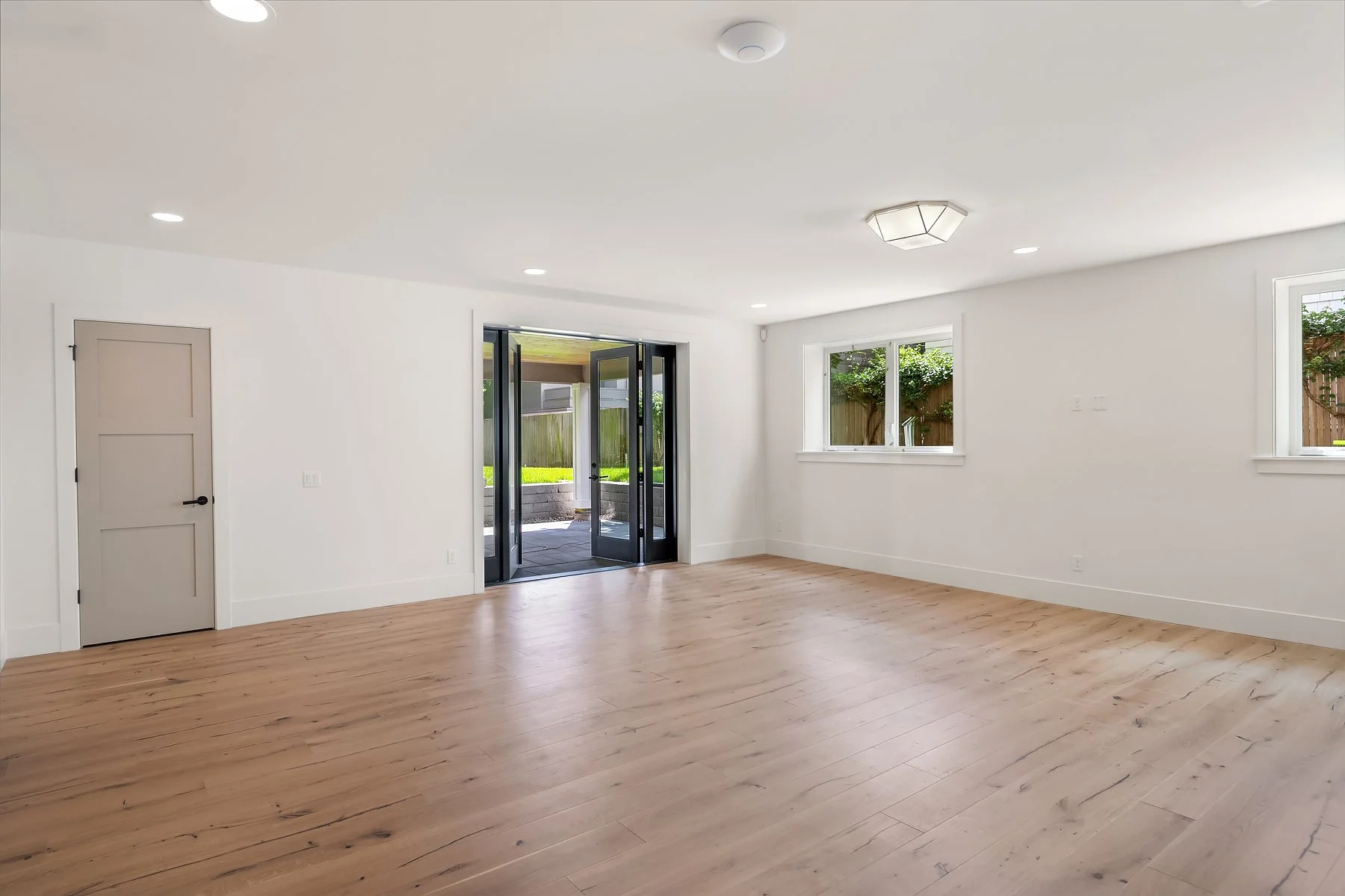 Empty living room with white walls, large windows, a glass sliding door, light wood flooring, and ceiling lights.