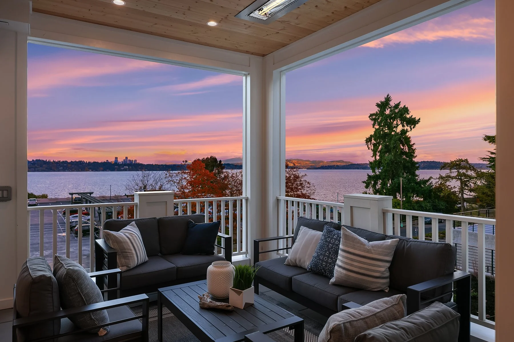 View of a balcony overlooking a lake at sunset with pink, purple, and orange sky, trees, and distant city skyline.