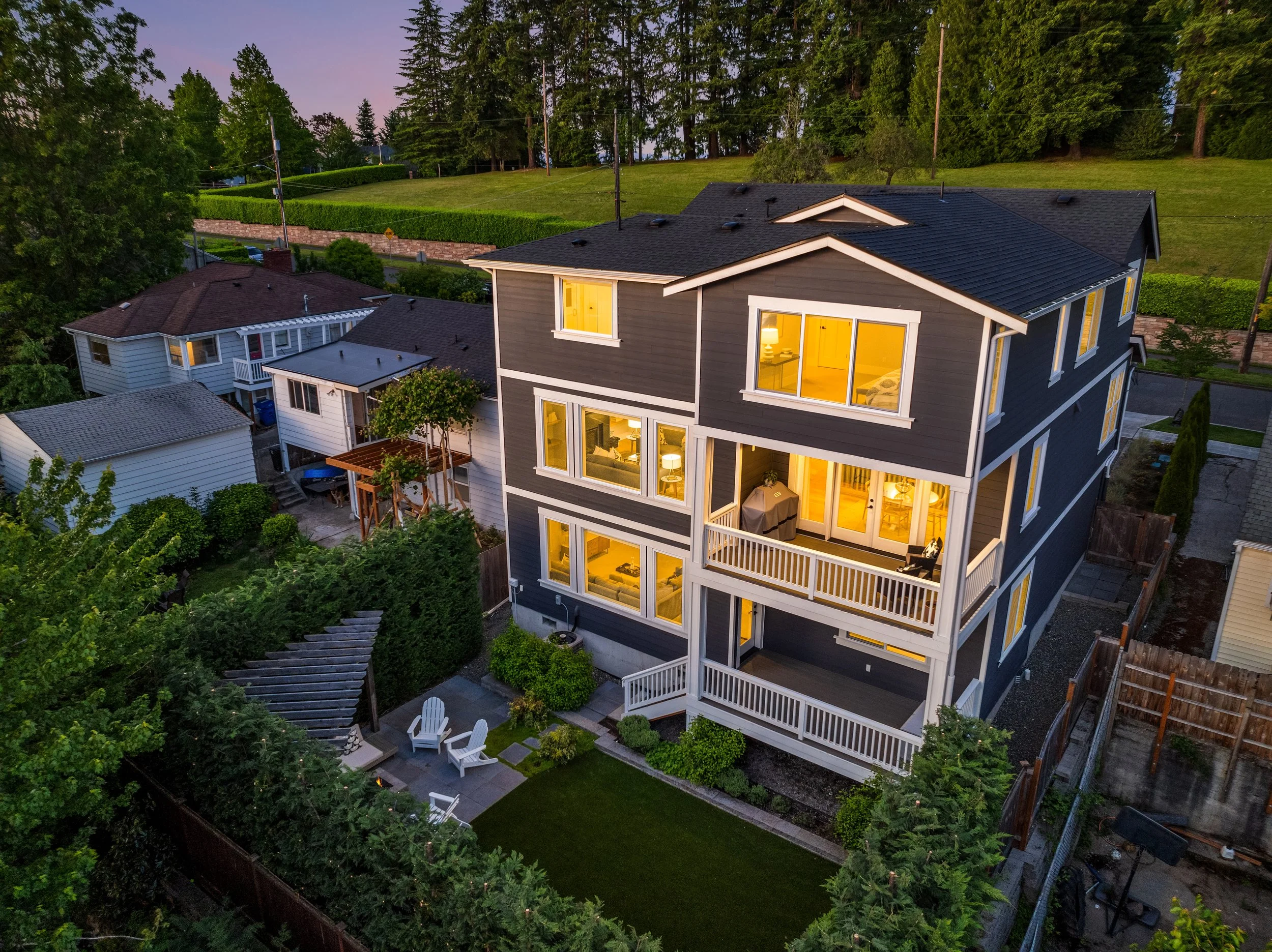 A three-story house with lit interior lights, featuring large windows and a multiple-level porch, surrounded by trees, neighboring houses, and a backyard with outdoor furniture at dusk.