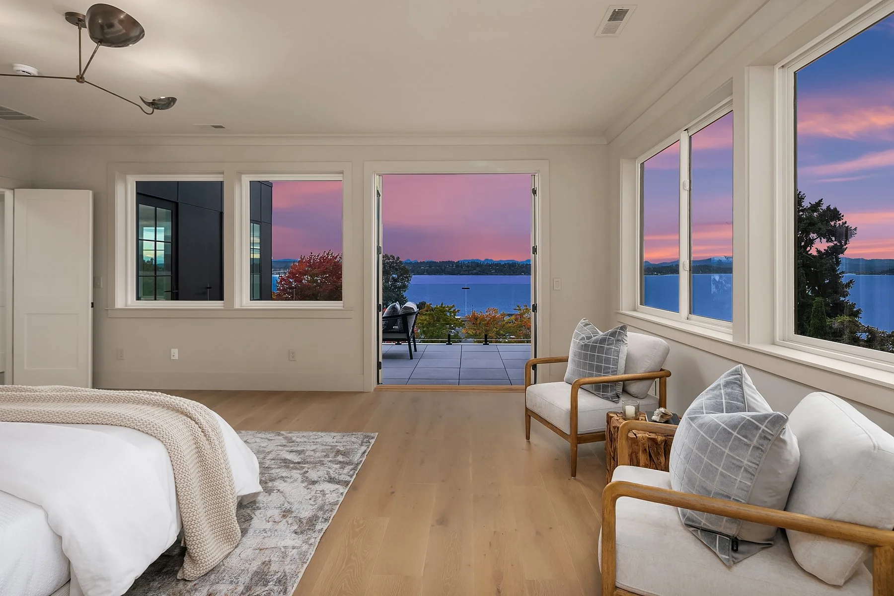 A bedroom with large windows and a door opening to a balcony overlooking a body of water and a colorful sunset sky. The room has a bed, two armchairs with pillows, and a small wooden side table.