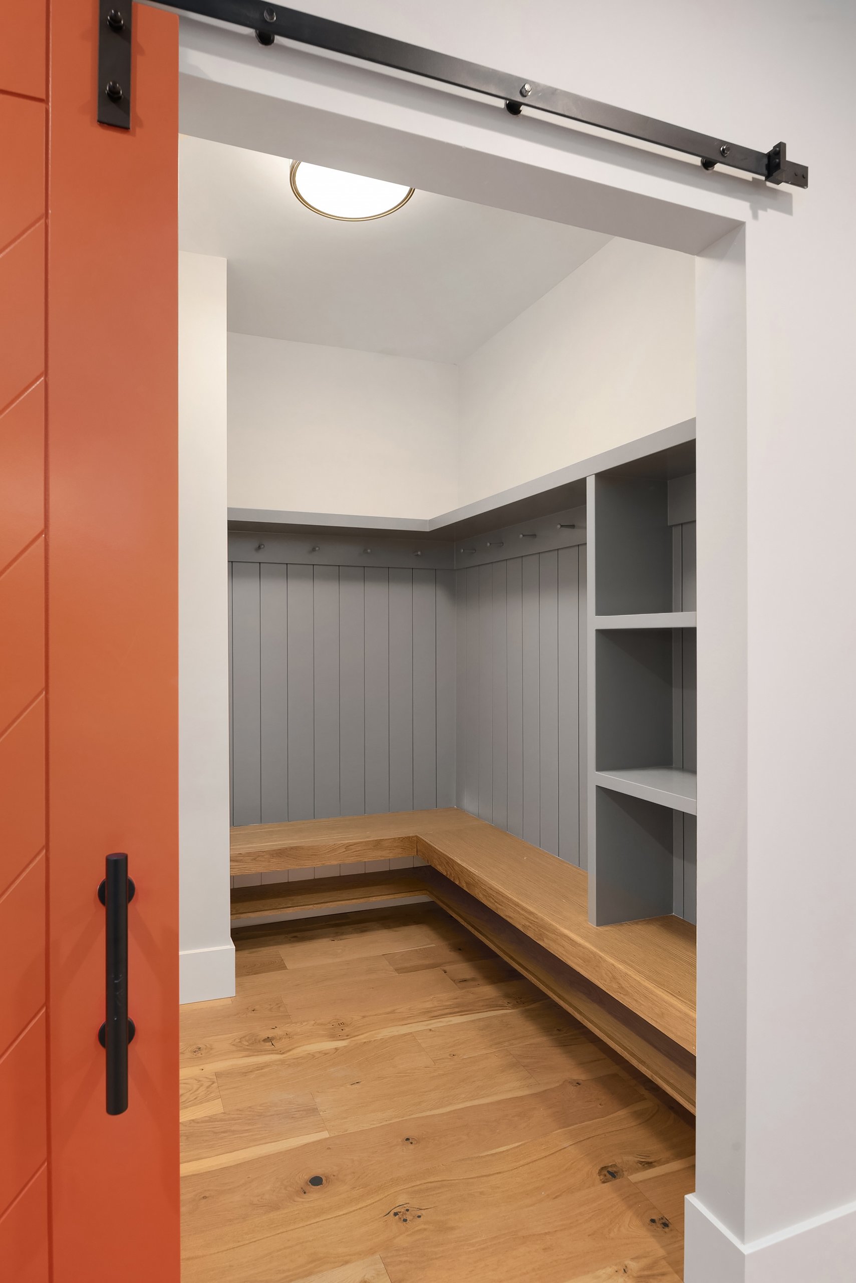 Interior of a small mudroom or closet with a wooden bench, shelves, gray beadboard wall panels, hardwood floors, and a white ceiling with a ceiling light. A sliding barn door with black hardware is partially visible on the left.