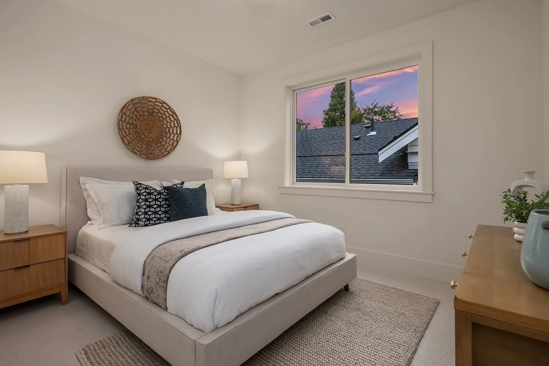 A cozy bedroom with a large white bed, nightstands with lamps, decorative pillows, a woven wall hanging, and a window showing a sunset sky and neighboring rooftops.