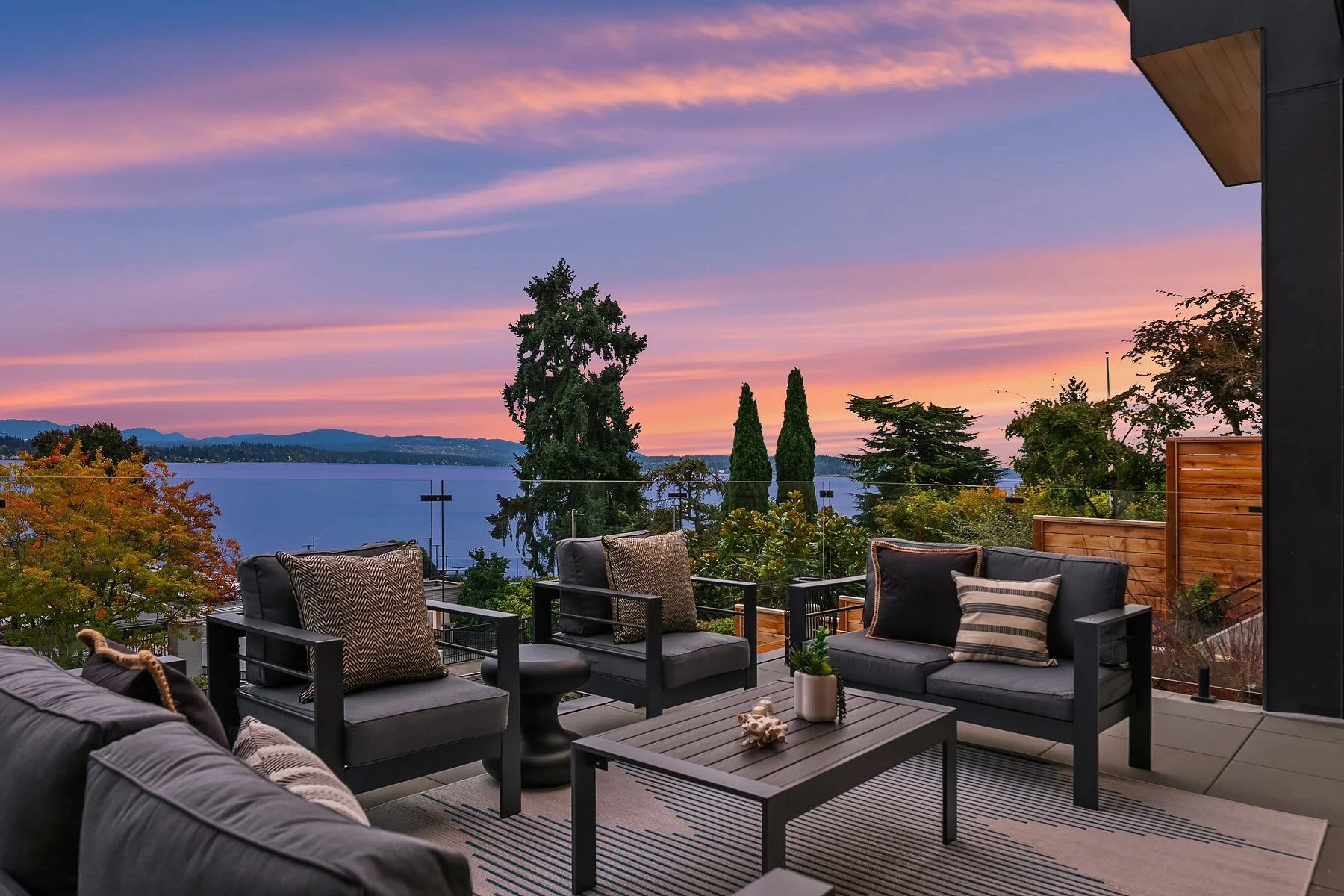 Outdoor patio furniture with black chairs and a coffee table on a balcony overlooking a lake and sunset sky with colorful clouds and trees in the background.