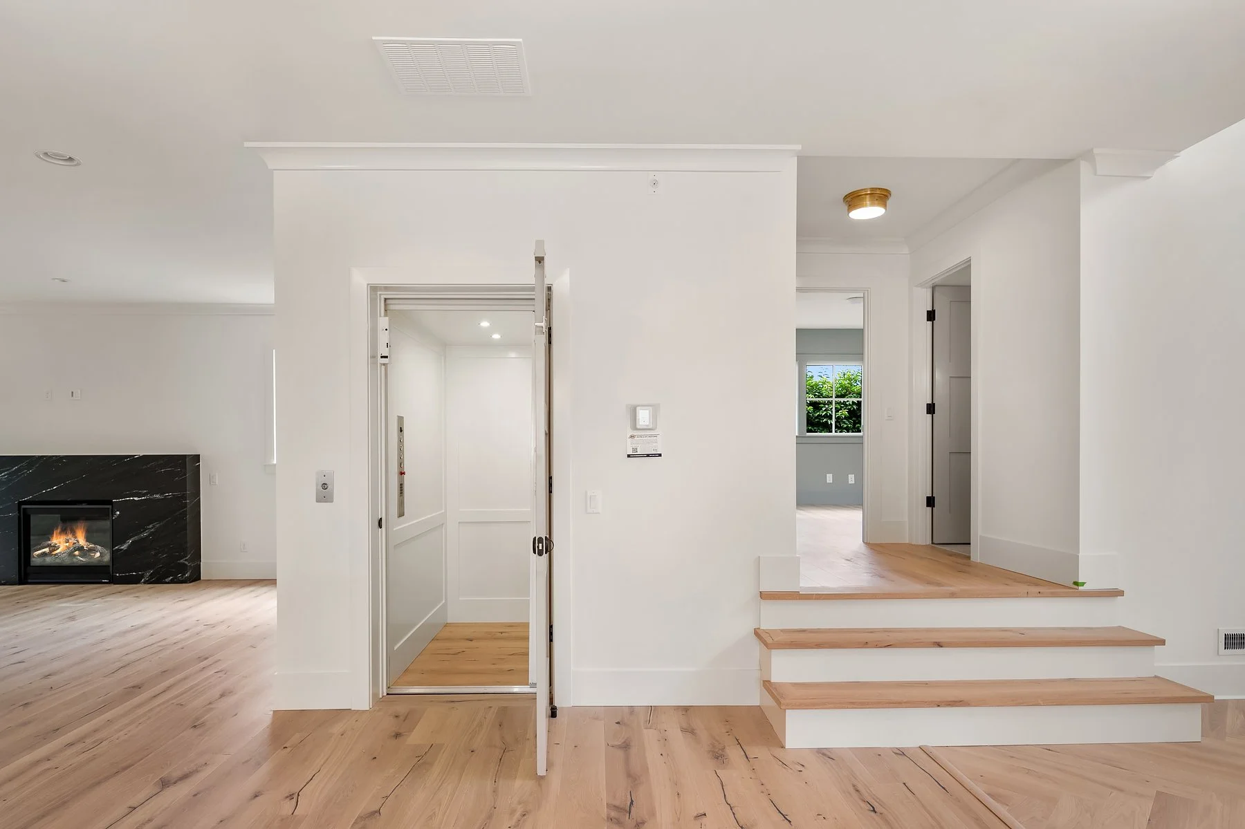 Interior view of a modern house with light-colored hardwood floors, white walls, stairs leading to a higher level, an elevator, and a glimpse of a room with a window showing greenery outside.