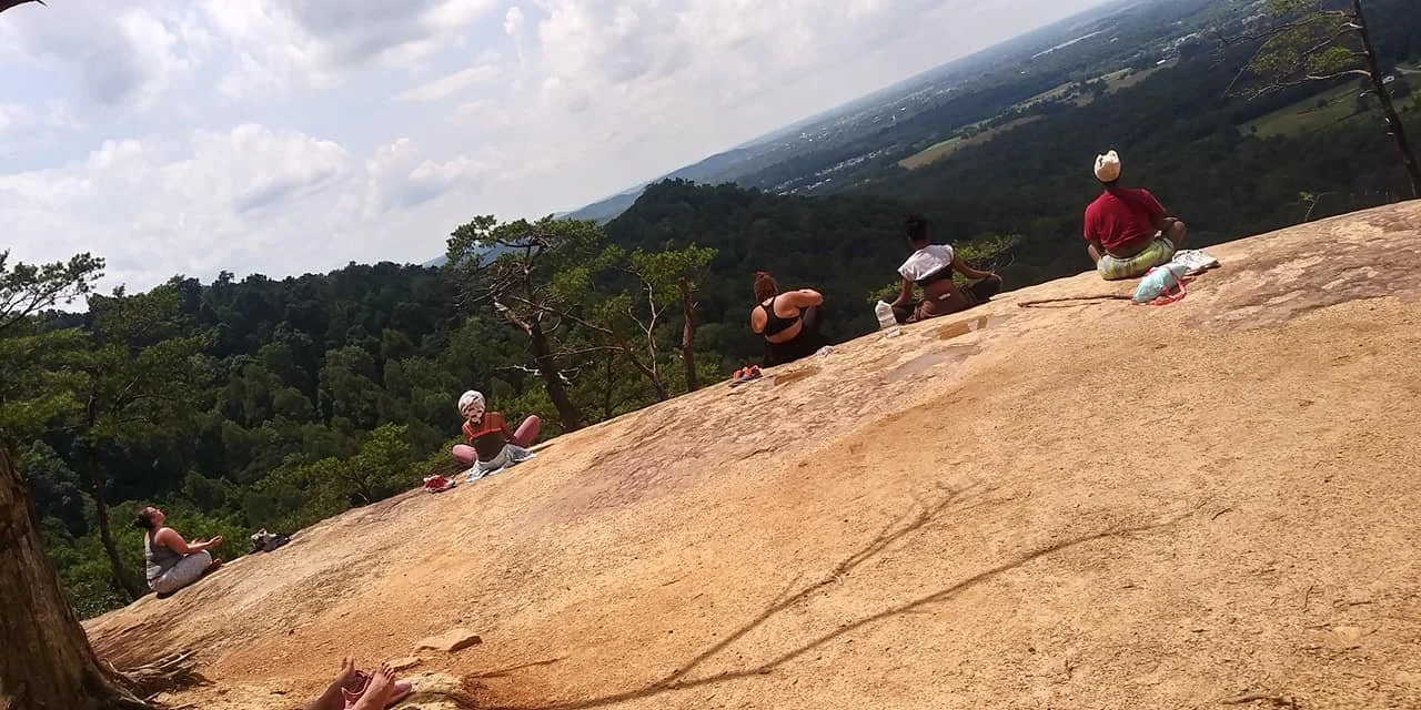 People practicing yoga or meditation on a large rock formation outdoors, with a forested valley and cloudy sky in the background.