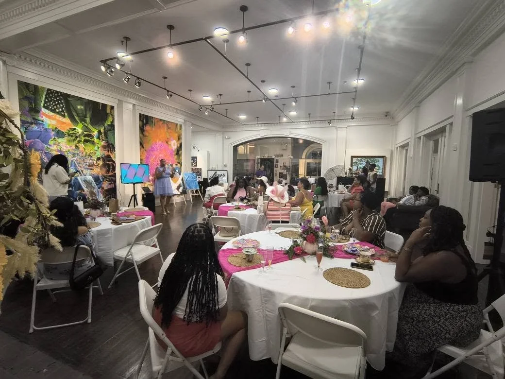 Women attending a presentation or celebration in a decorated event space with round tables, floral centerpieces, and large colorful artwork on the walls.