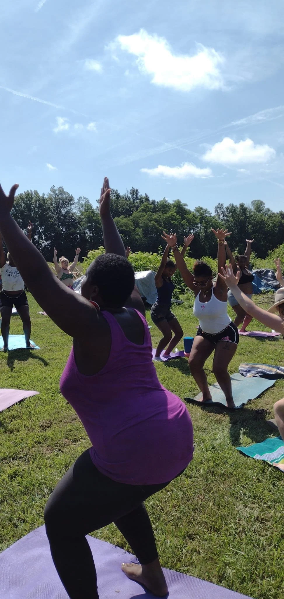 Group of women practicing yoga outdoors on grassy field under blue sky with scattered clouds, surrounded by trees.