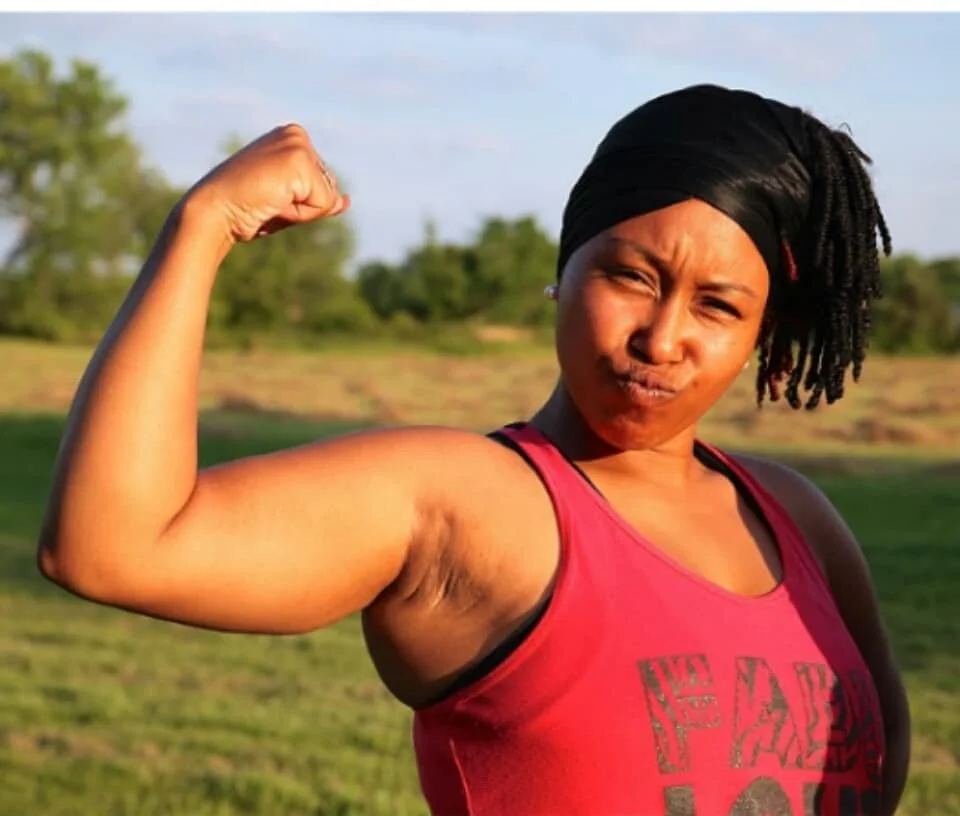 Woman flexing her right arm outdoors on a grassy field, wearing a red tank top and black headwrap, with trees and a blue sky in the background.