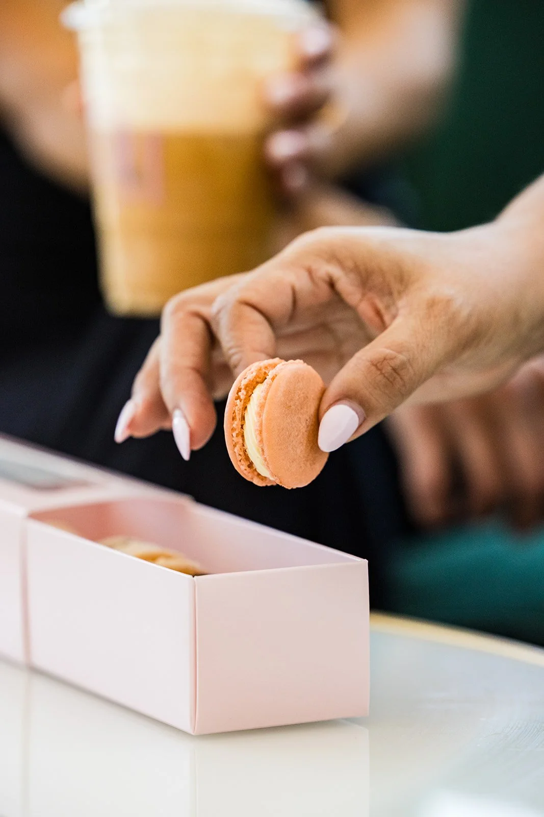 Person holding a pink macaroon cookie over a pink box, with someone in the background drinking a iced coffee — handcrafted macarons and desserts at Decadent Macaron café in Gilbert, Arizona