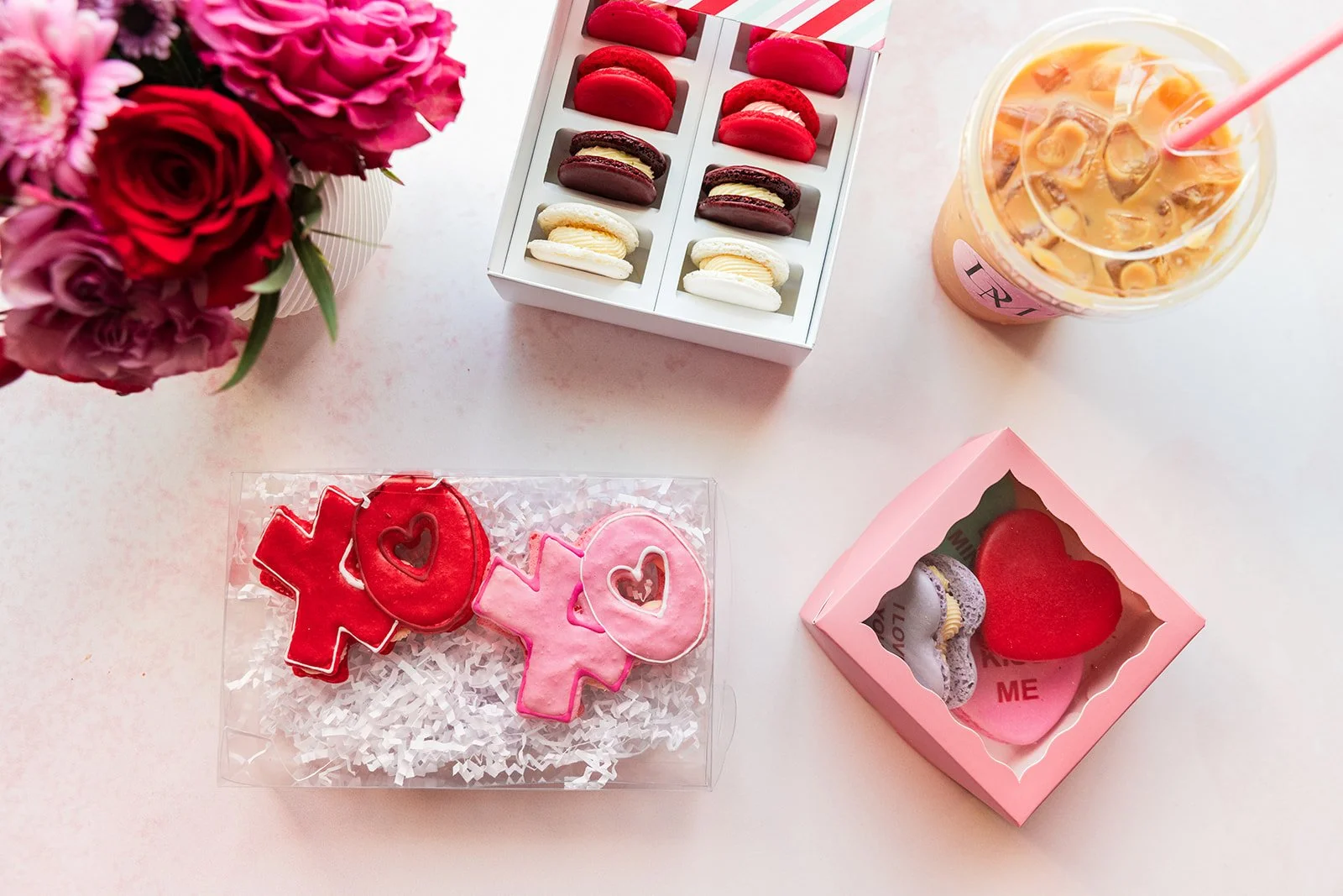 A bouquet of pink, red, and purple flowers, a box of assorted French macarons, a pink drink with ice and a straw, and heart-shaped cookies with pink, red, and purple icing on a light pink surface — handcrafted macarons and desserts at Decadent Macaro