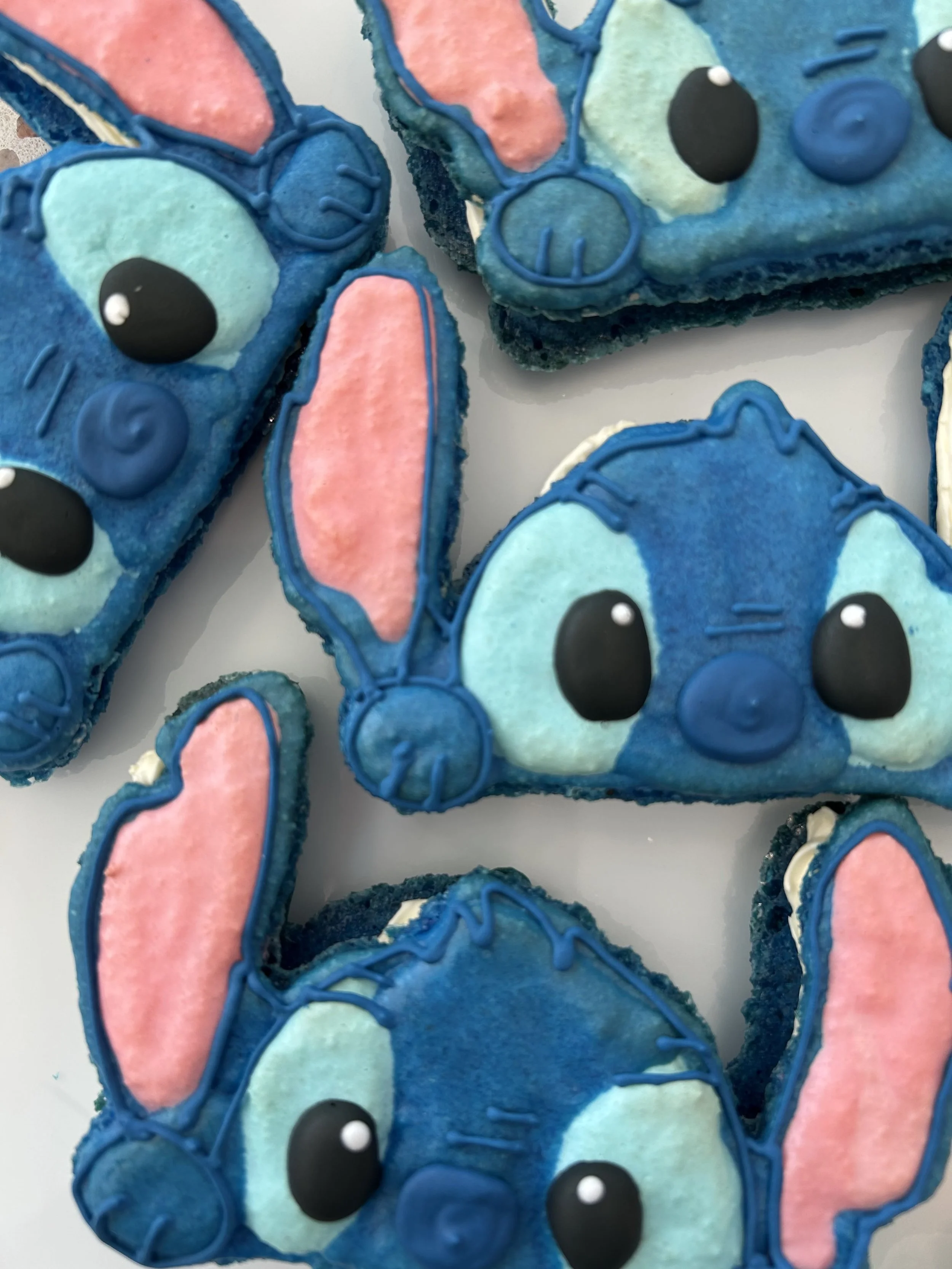 Decorated cookies shaped like Disney's Stitch with big eyes, pink inner ears, and blue icing, arranged on a white surface — custom dessert catering and events by Decadent Macaron in Gilbert, Arizona